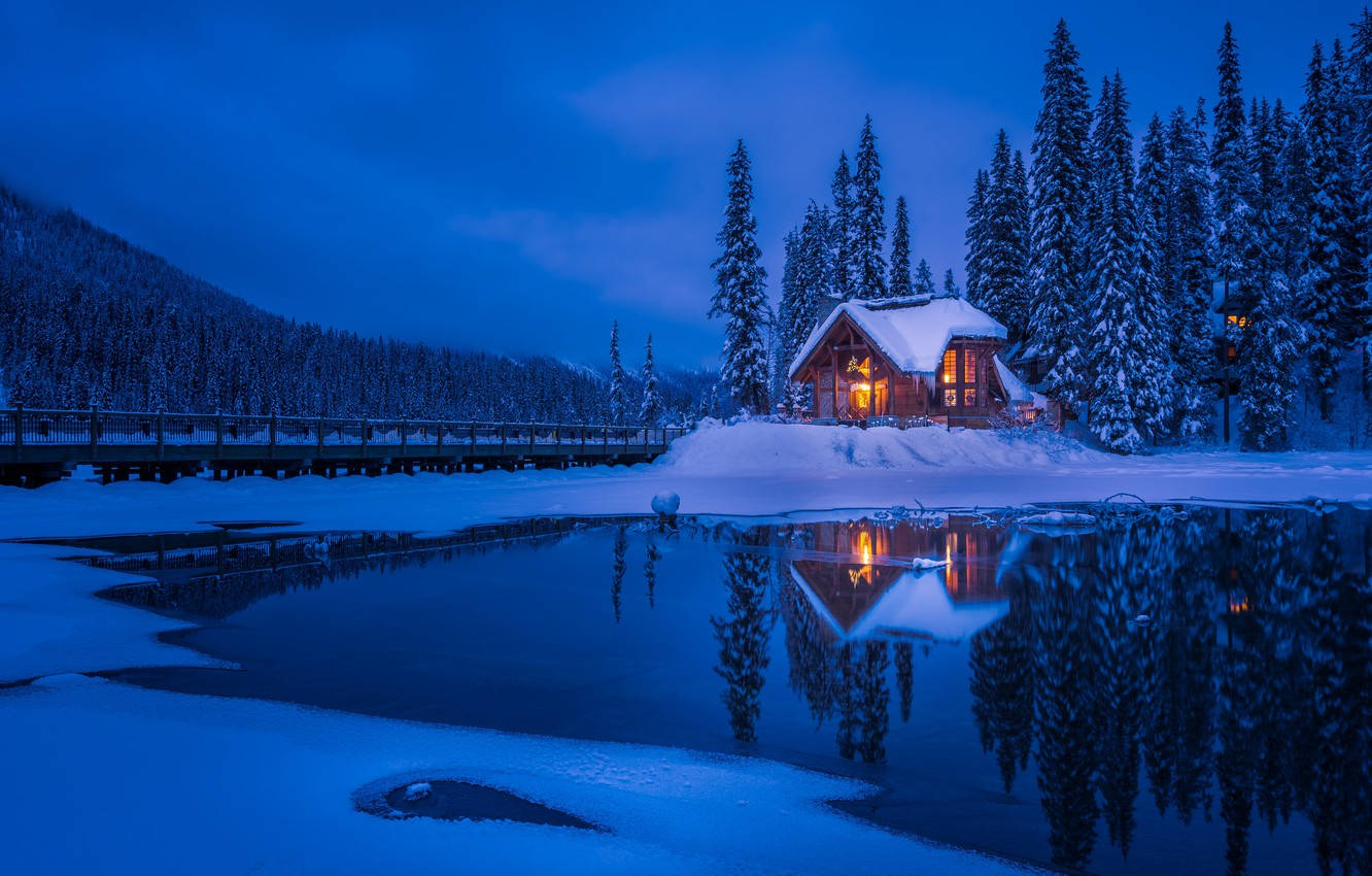 Winter House 桌布 壁紙 — 一間舒適的木屋在暮色中散發溫暖燈光，映襯著白雪覆蓋的松樹，倒影在結冰的湖面上閃爍。