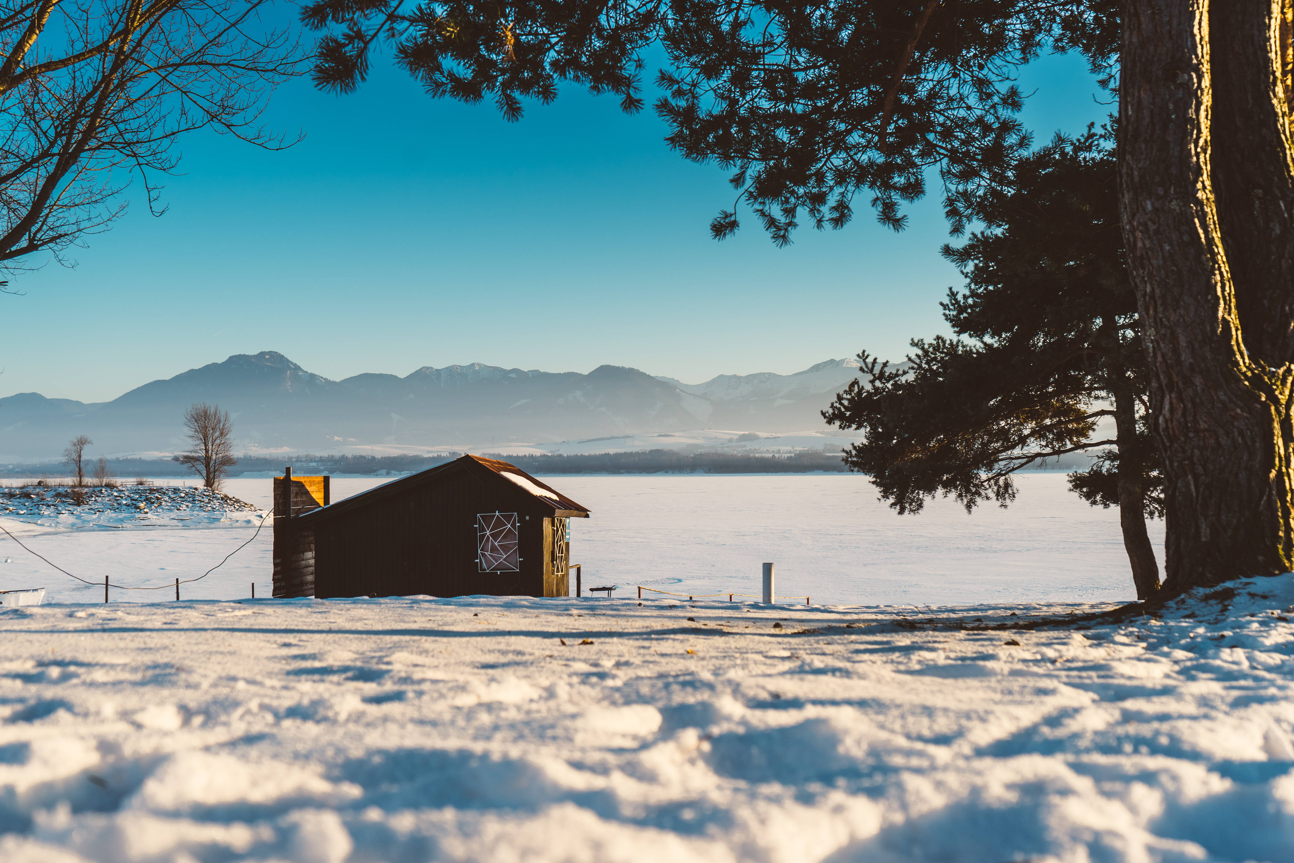 Slovakia 桌布 壁紙 — 一間舒適的木屋坐落在白雪皚皚的冬季景觀中，遠處群山環繞，清澈的藍天下松樹成蔭。