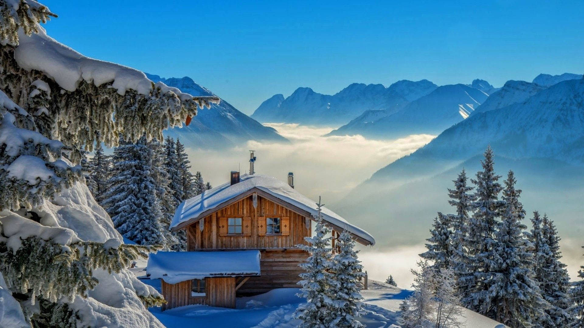 Winter House 桌布 壁紙 — 一間舒適的木屋坐落在白雪覆蓋的松樹林中，霧氣繚繞的山巒在雲海之上聳立，天空清澈湛藍。