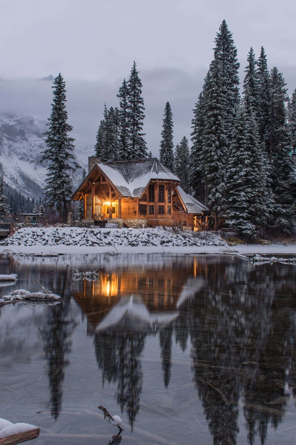 Cozy Winter Cabin 桌布 壁紙 — 一間舒適的木屋坐落在白雪覆蓋的松樹之間，在暮色中溫暖地倒映在平靜的冰湖上。