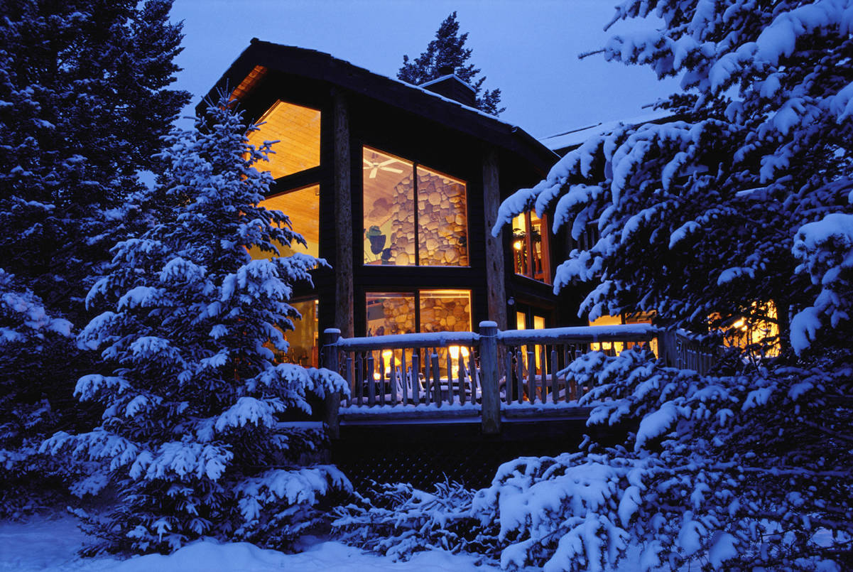 Winter House 桌布 壁紙 — 一間舒適的木屋坐落在白雪覆蓋的松樹之間，在暮色天空的映襯下從內部散發出溫暖的光芒。