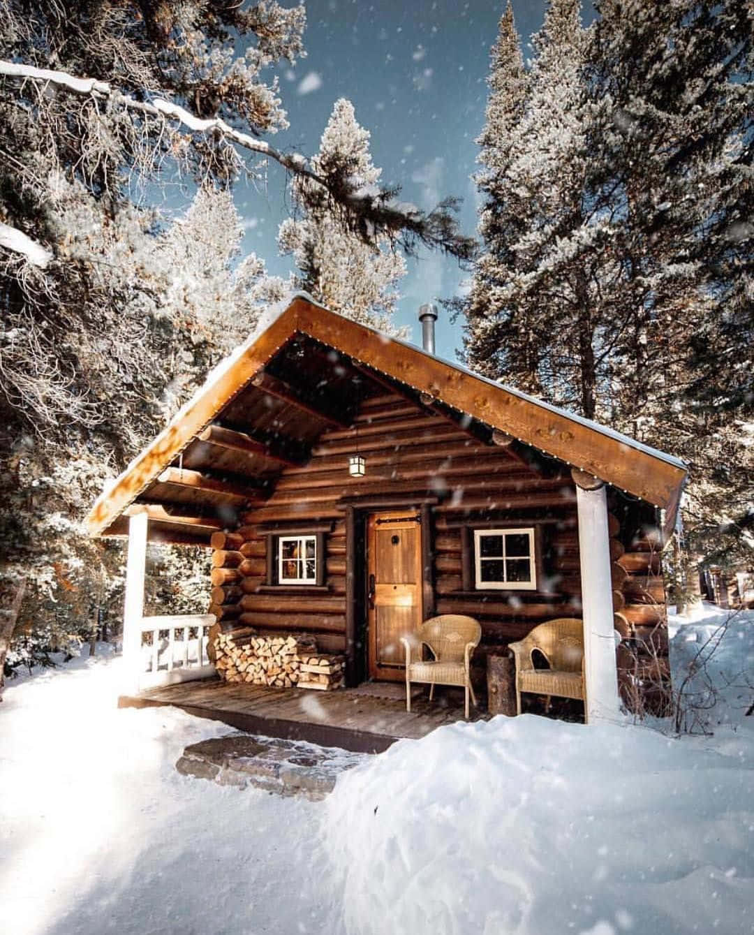 Log Cabin 桌布 壁紙 — 一間舒適的木屋坐落在白雪覆蓋的松樹林中，天空湛藍，雪花飄落。