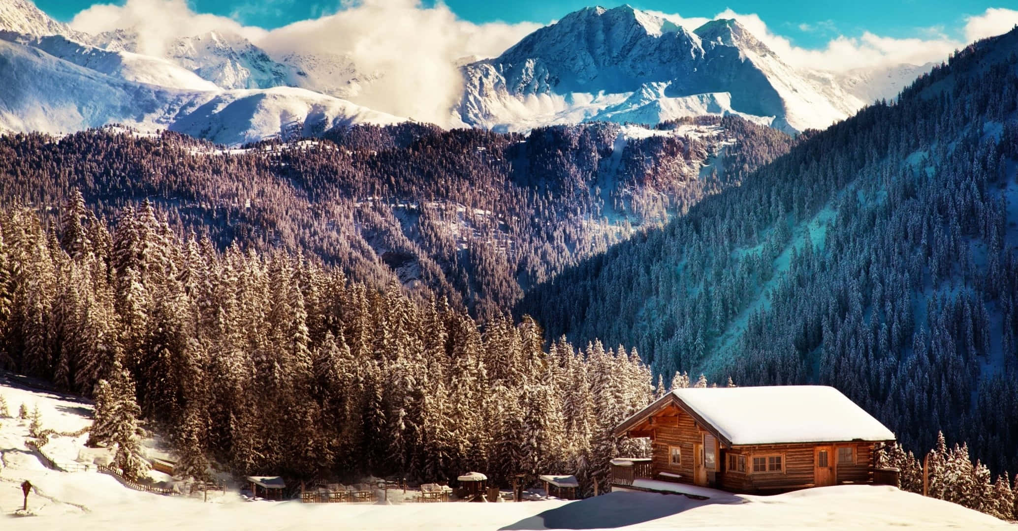 山景 桌布 壁紙 — 一間舒適的木屋坐落在白雪覆蓋的松樹林中，背景是雄偉的雪山，上方是明亮的藍天。