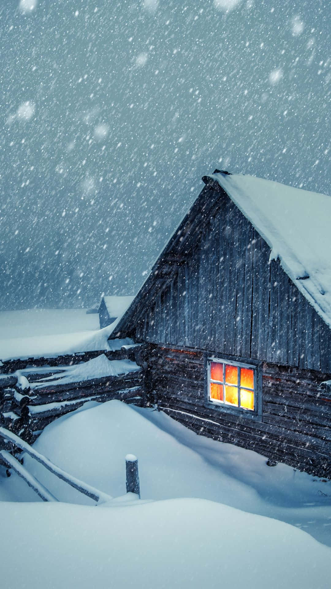 Snow Falling 桌布 壁紙 — 一間舒適的木屋隱藏在暴風雪中，溫暖發光的窗戶劃破了冰冷的藍色冬日場景。
