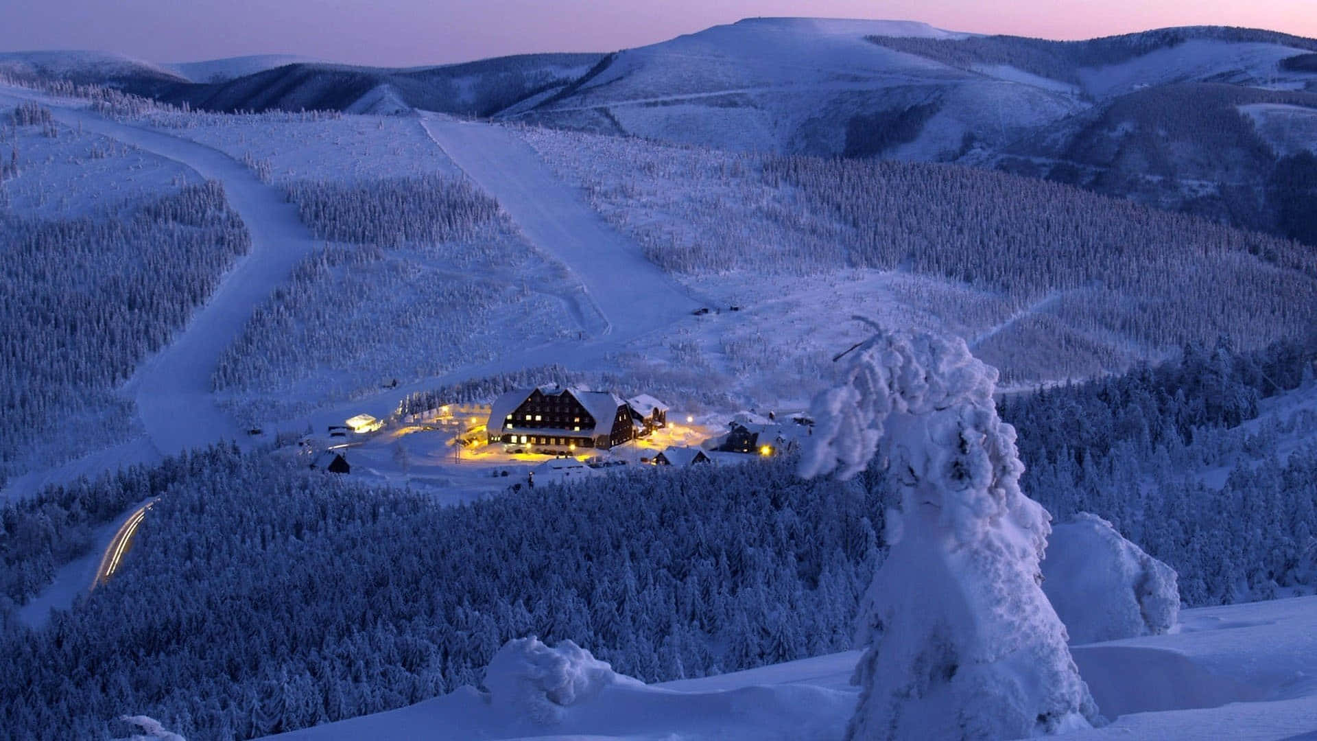 Ski Mountain 桌布 壁紙 — 一間舒適的滑雪小屋在暮色中溫暖地發光，映襯著白雪覆蓋的山景，燈火通明的滑雪道蜿蜒穿過森林。