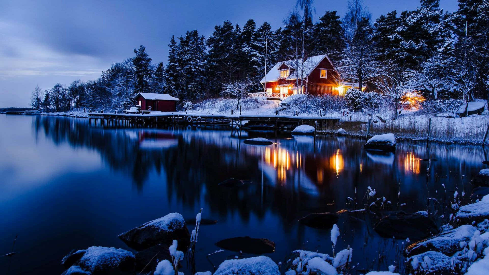 1920x1080 Winter 桌布 壁紙 — 一間舒適的紅色小屋，在暮色天空下，溫暖的燈光在白雪覆蓋的湖邊閃爍。