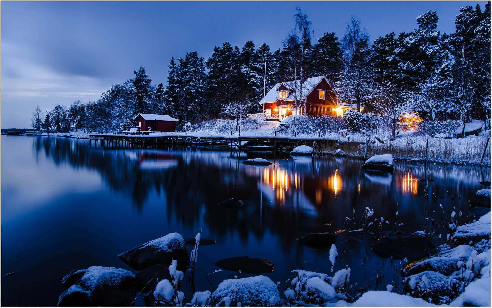 Best Winter 桌布 壁紙 — 一間舒適的紅色小屋在暮色中散發溫暖燈光，映襯著白雪覆蓋的森林，其倒影在平靜的冰水中閃爍。