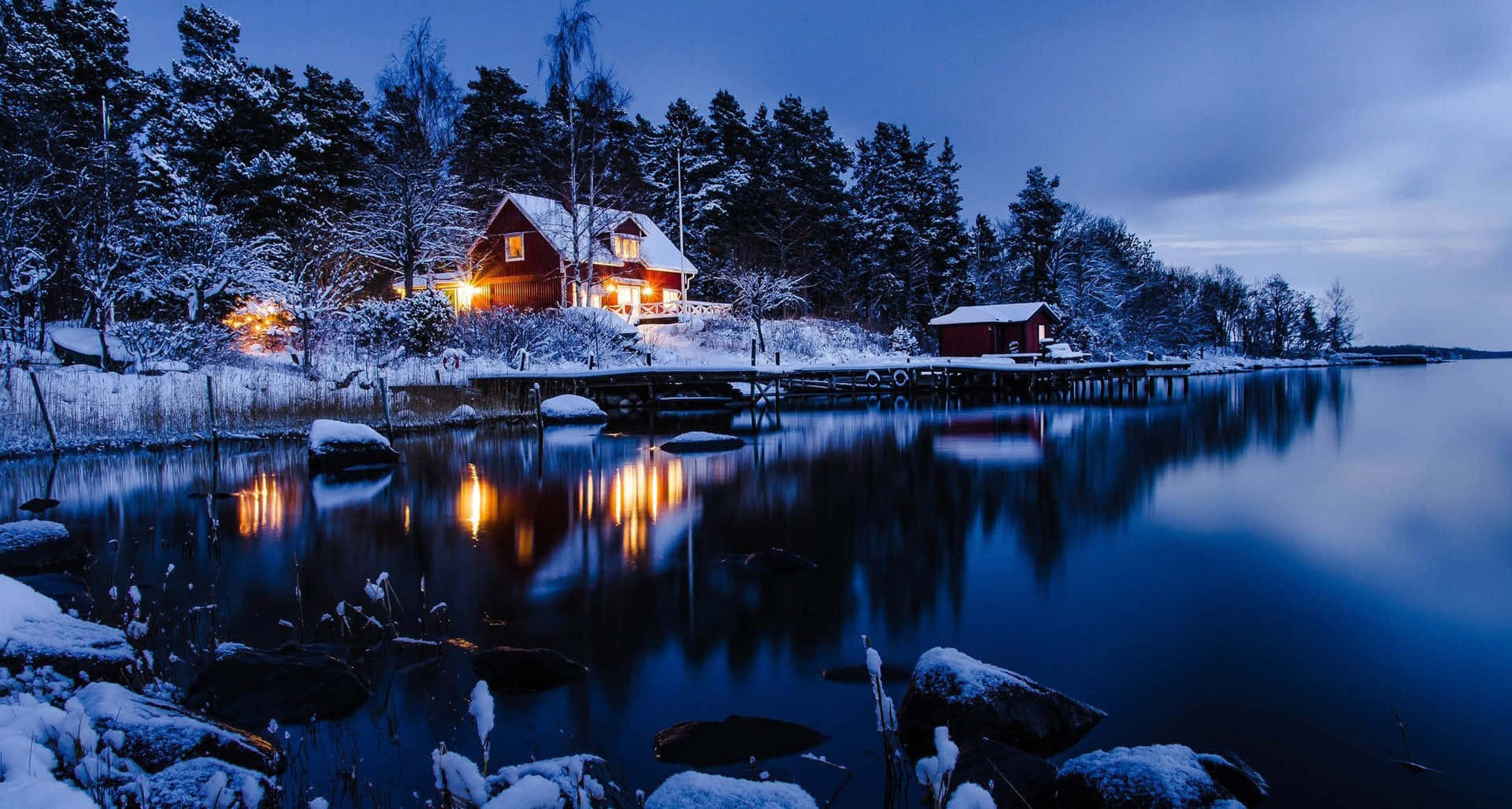 Winter Scenery Desktop 桌布 壁紙 — 一間舒適的紅色小屋在雪地黃昏的海岸線上溫暖地發光，燈光倒映在平靜冰冷的水面上。