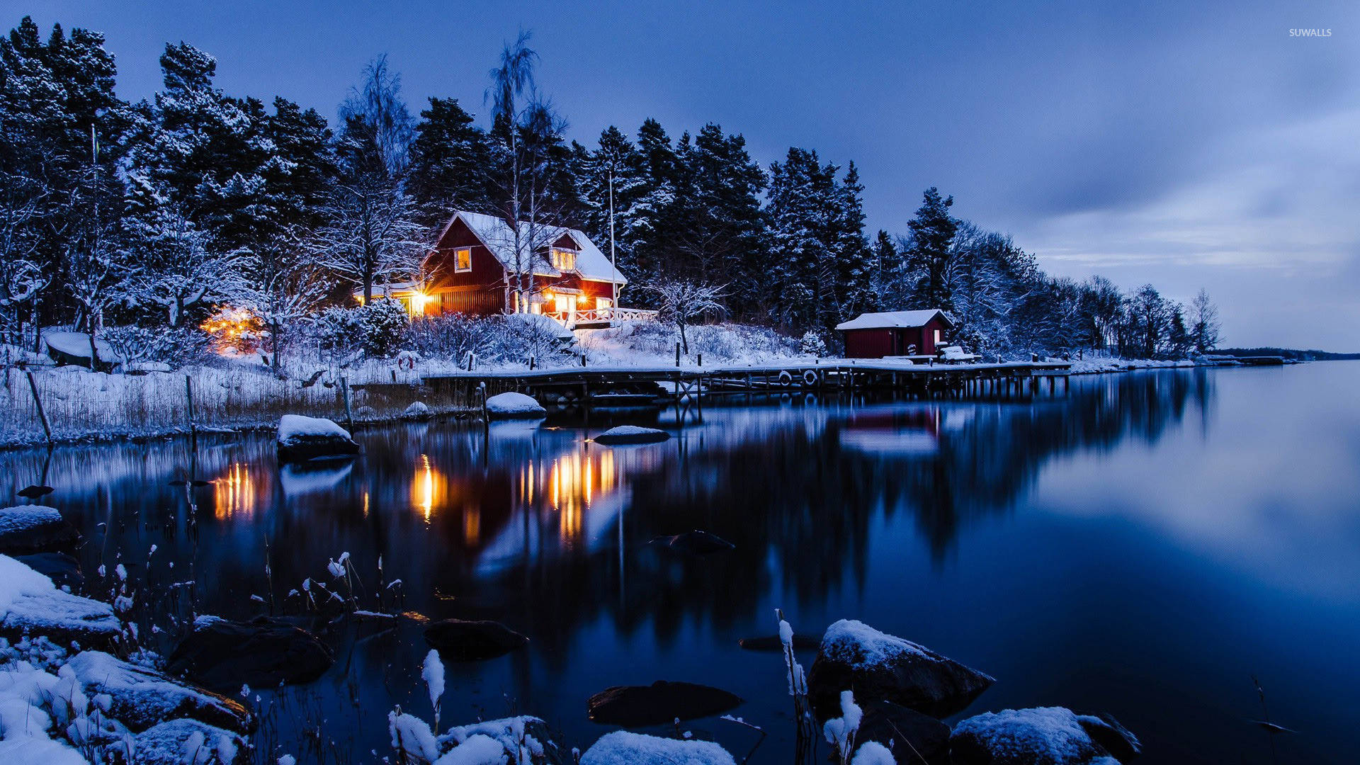 Winter Landscape 桌布 壁紙 — 一間舒適的紅色小屋在黃昏時分，在白雪覆蓋的冬日湖邊溫暖地發光，平靜的水面上閃爍著倒影。