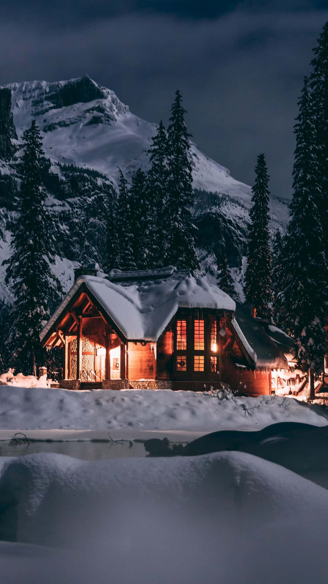 Cute Winter Phone 桌布 壁紙 — 一間舒適的被雪覆蓋的小屋在黑暗的山景背景下，在雪夜的天空下溫暖地發光。