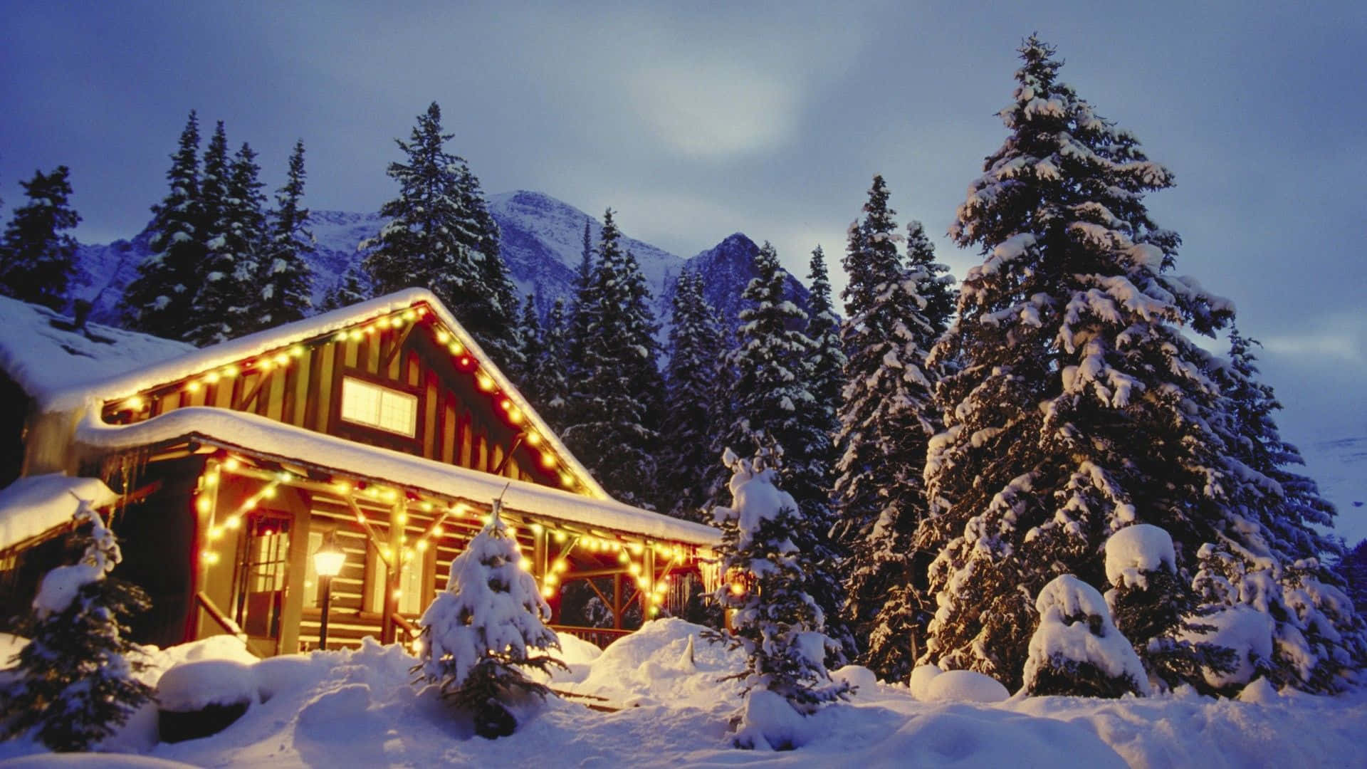 Cozy Winter Cabin 桌布 壁紙 — 一間舒適的雪覆蓋小屋，在暮色山景背景下，用串燈溫暖地發光。