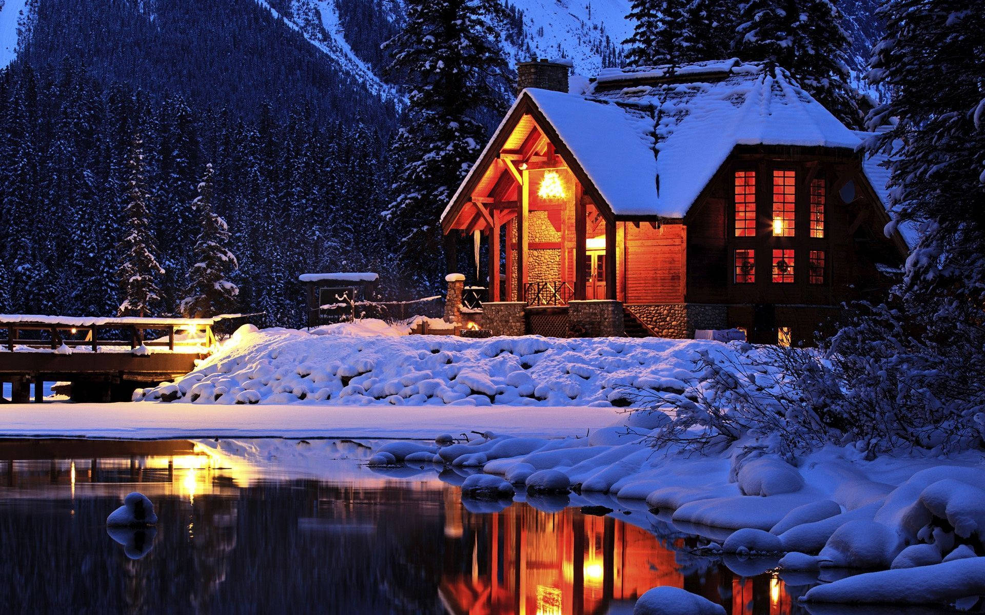 old 桌布 壁紙 — 一間舒適的雪覆蓋小屋在暮色山巒背景下溫暖地發光，其燈光反射在結冰的湖面上。