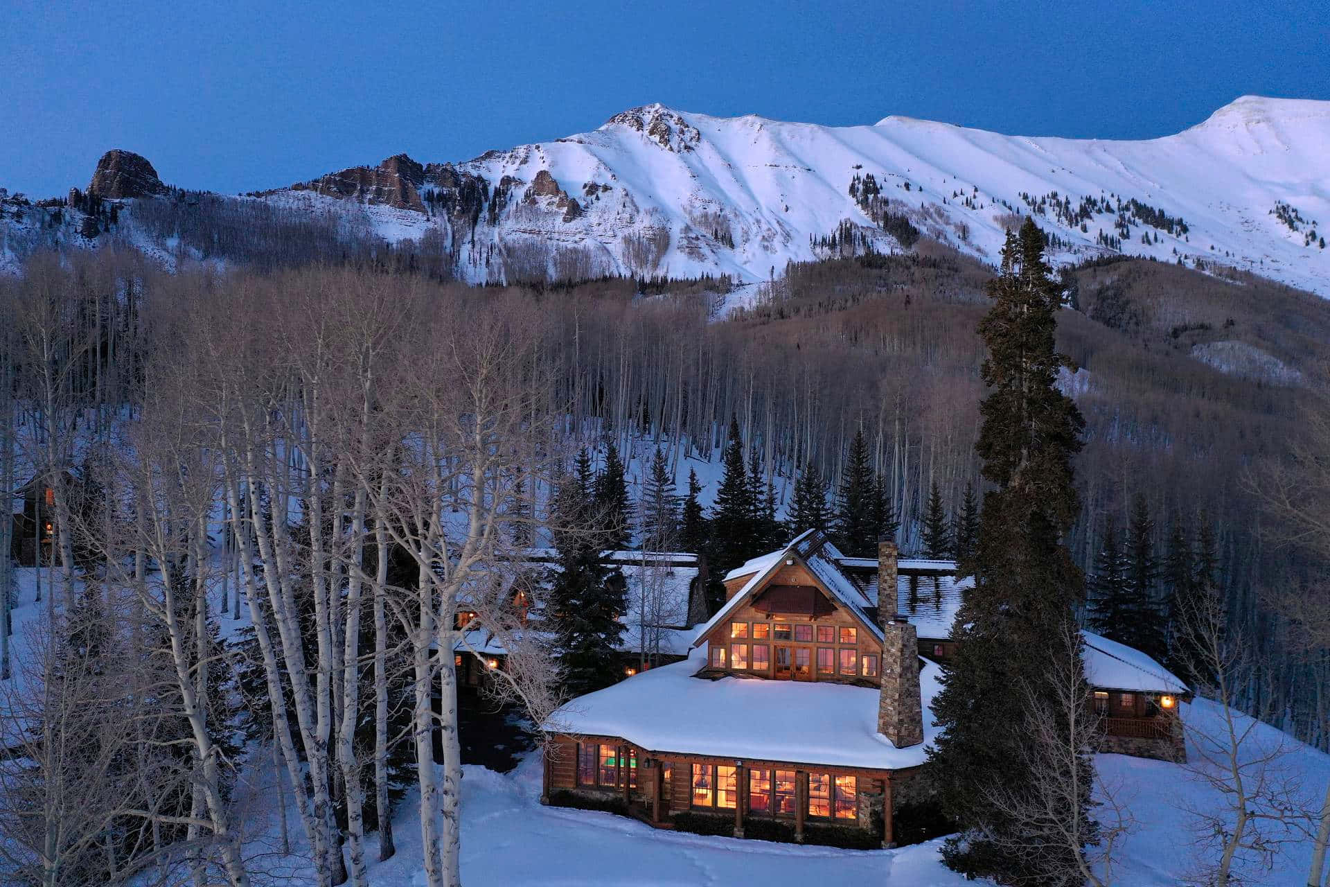 Colorado Mountain 桌布 壁紙 — 一間舒適的雪覆蓋小屋在暮色山景和光禿的白楊樹襯托下溫暖地發光。