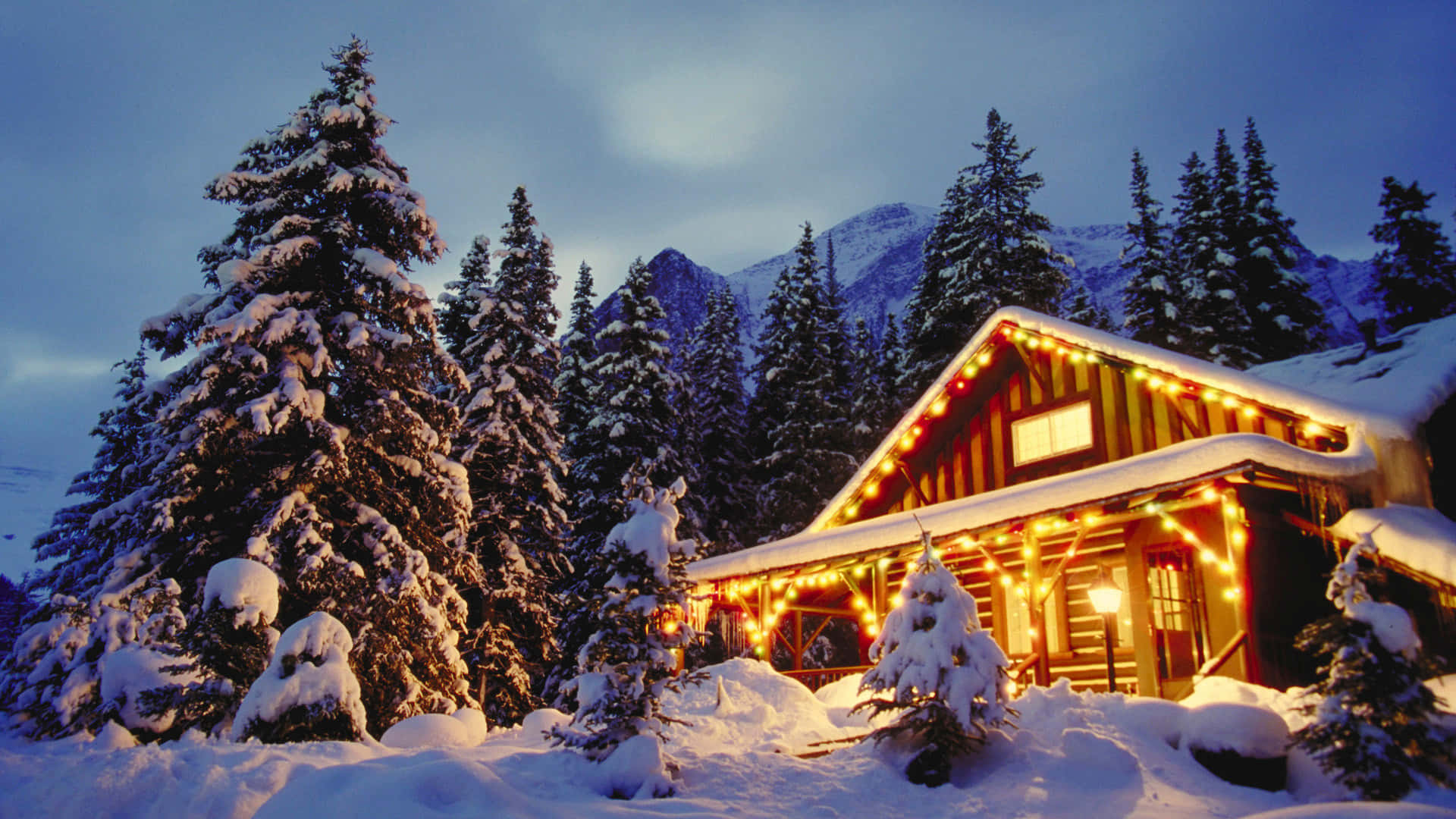 Christmas Snow 桌布 壁紙 — 一間舒適的雪覆蓋小屋在暮色山景背景下，用串燈溫暖地發光。