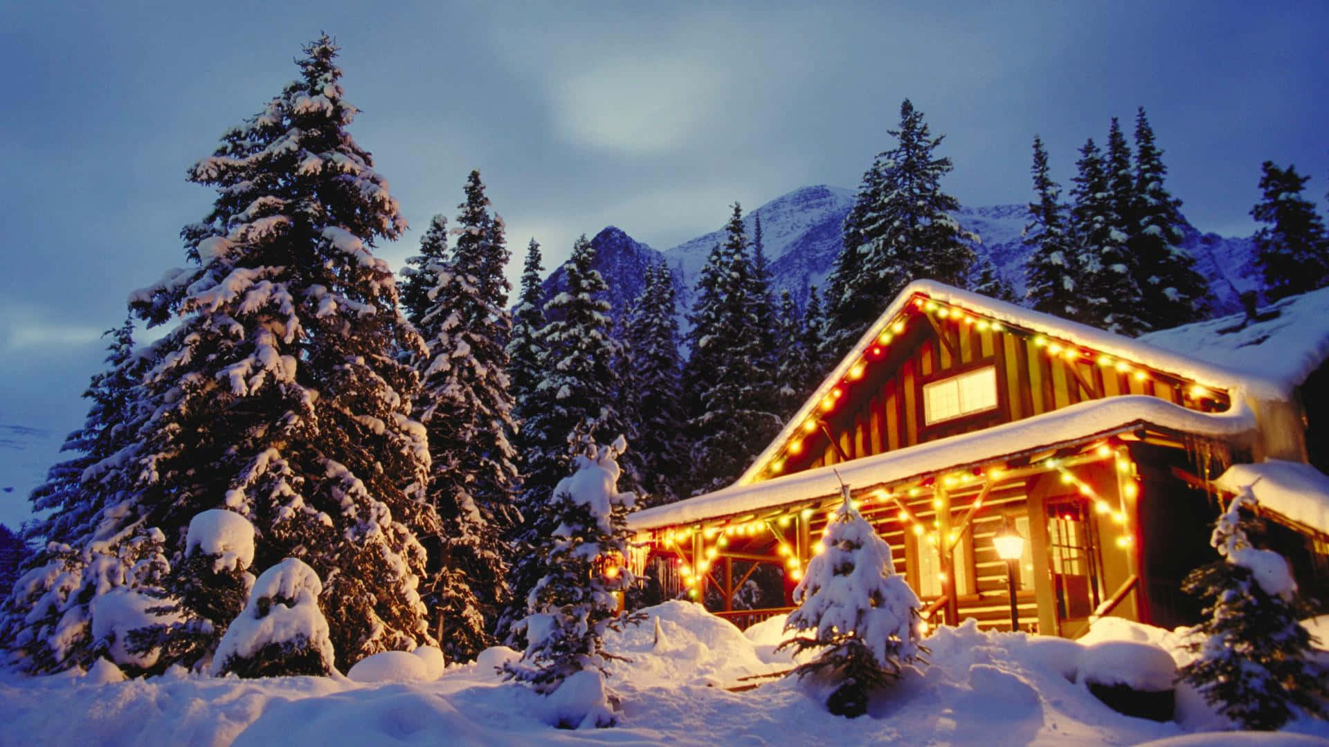 Winter Holiday 桌布 壁紙 — 一間舒適的雪覆蓋小屋在暮色山景背景下，用串燈溫暖地發光。