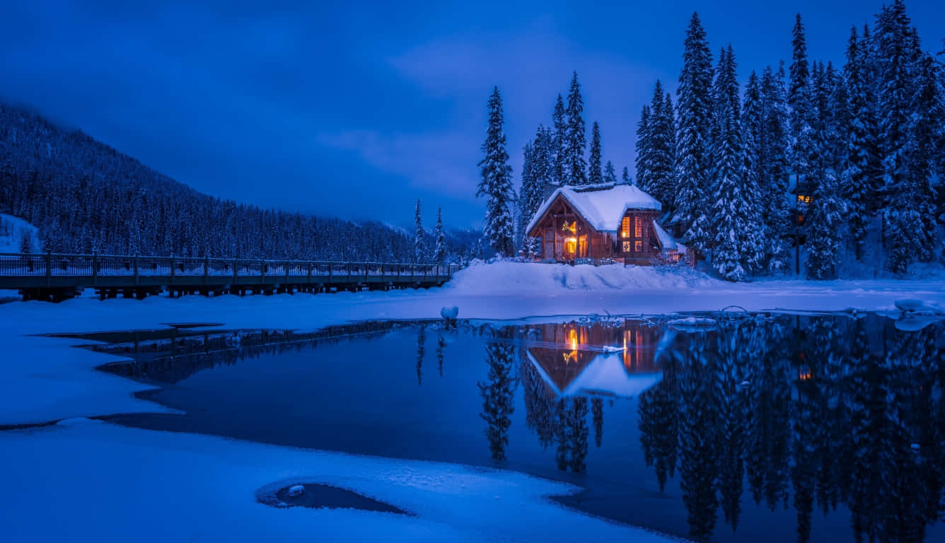 Winter Laptop 桌布 壁紙 — 一間舒適的雪覆蓋小屋在深藍色的冬日暮色中溫暖地發光，其倒影在一個部分結冰、被松樹環繞的湖中閃爍。