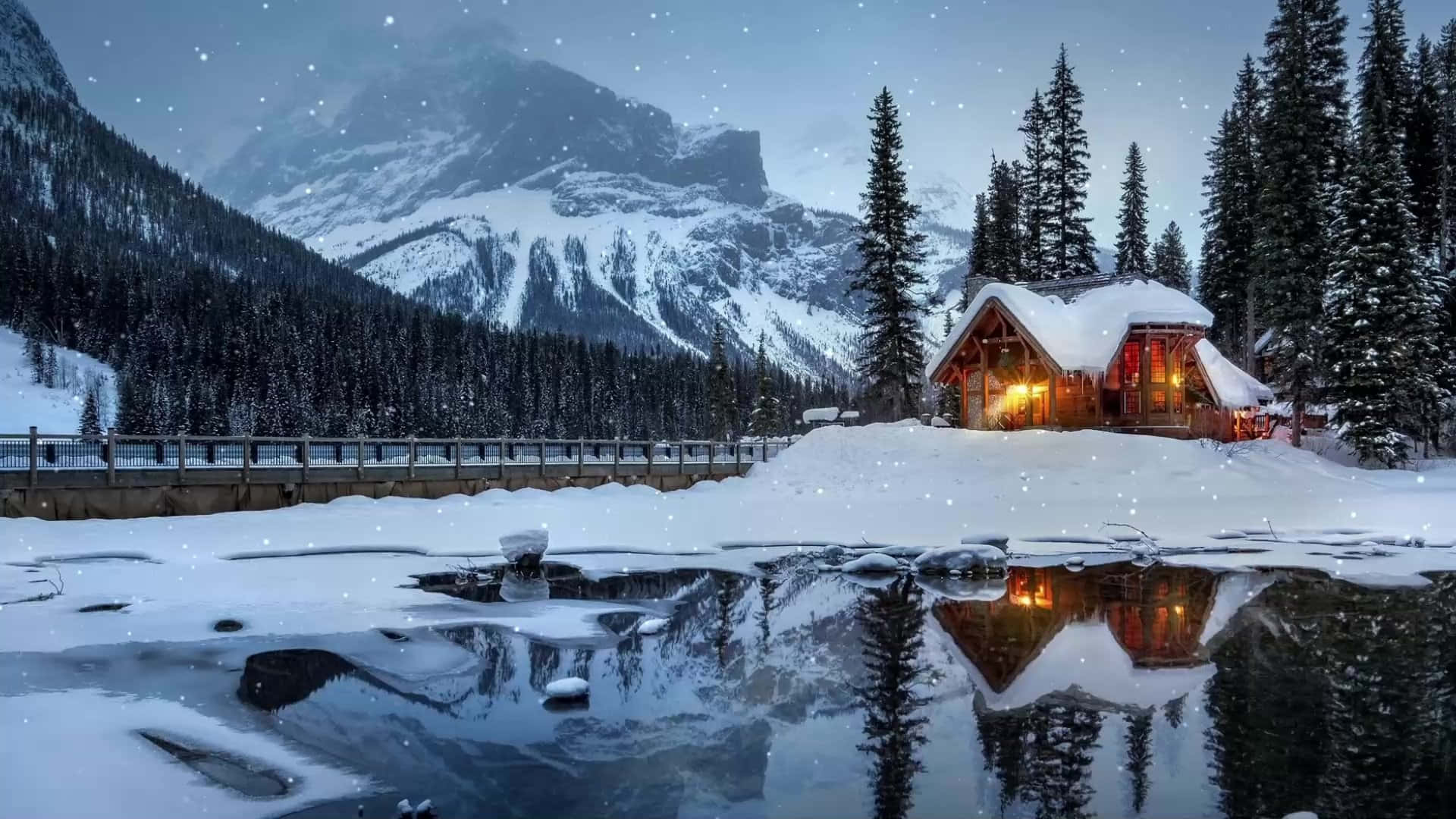 Cozy Winter Cabin 桌布 壁紙 — 一間舒適的雪覆蓋小屋在雄偉的雪山前溫暖地發光，雪花飄落在反射的冰凍湖面上。