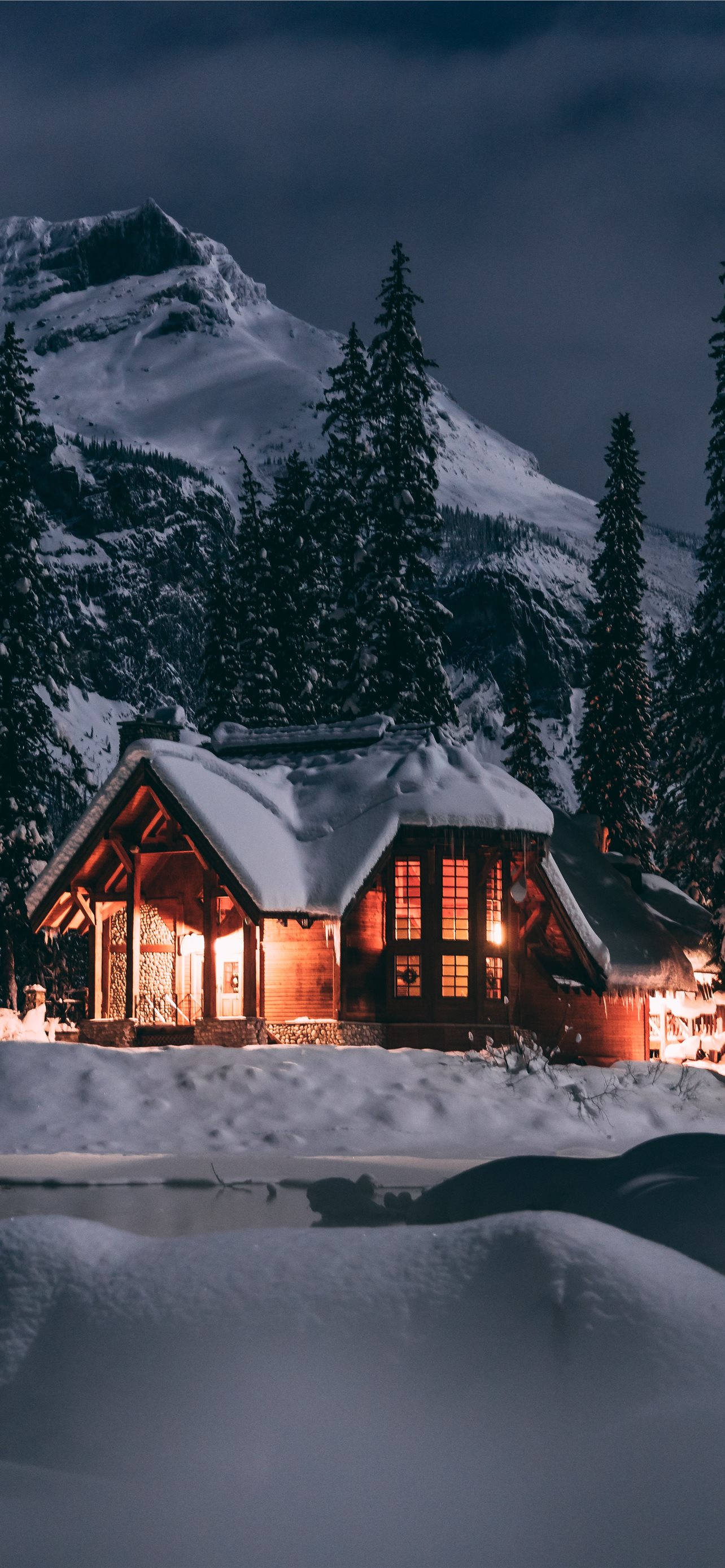 冬季 iPhone 壁紙 桌布 壁紙 — 一間舒適的雪覆蓋小屋在黑暗的山景背景下，在雪夜天空下溫暖地發光。