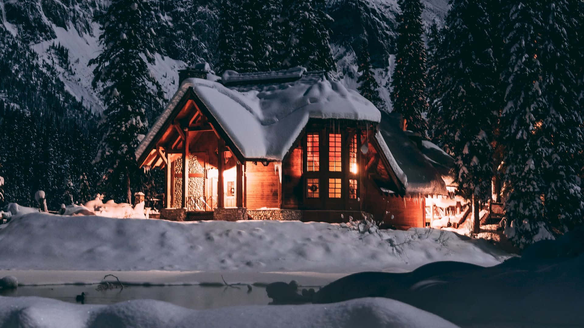 1920x1080 Winter 桌布 壁紙 — 一間舒適的雪覆蓋木屋在夜晚溫暖地發光，坐落在山區冬季森林中積雪的松樹之間。