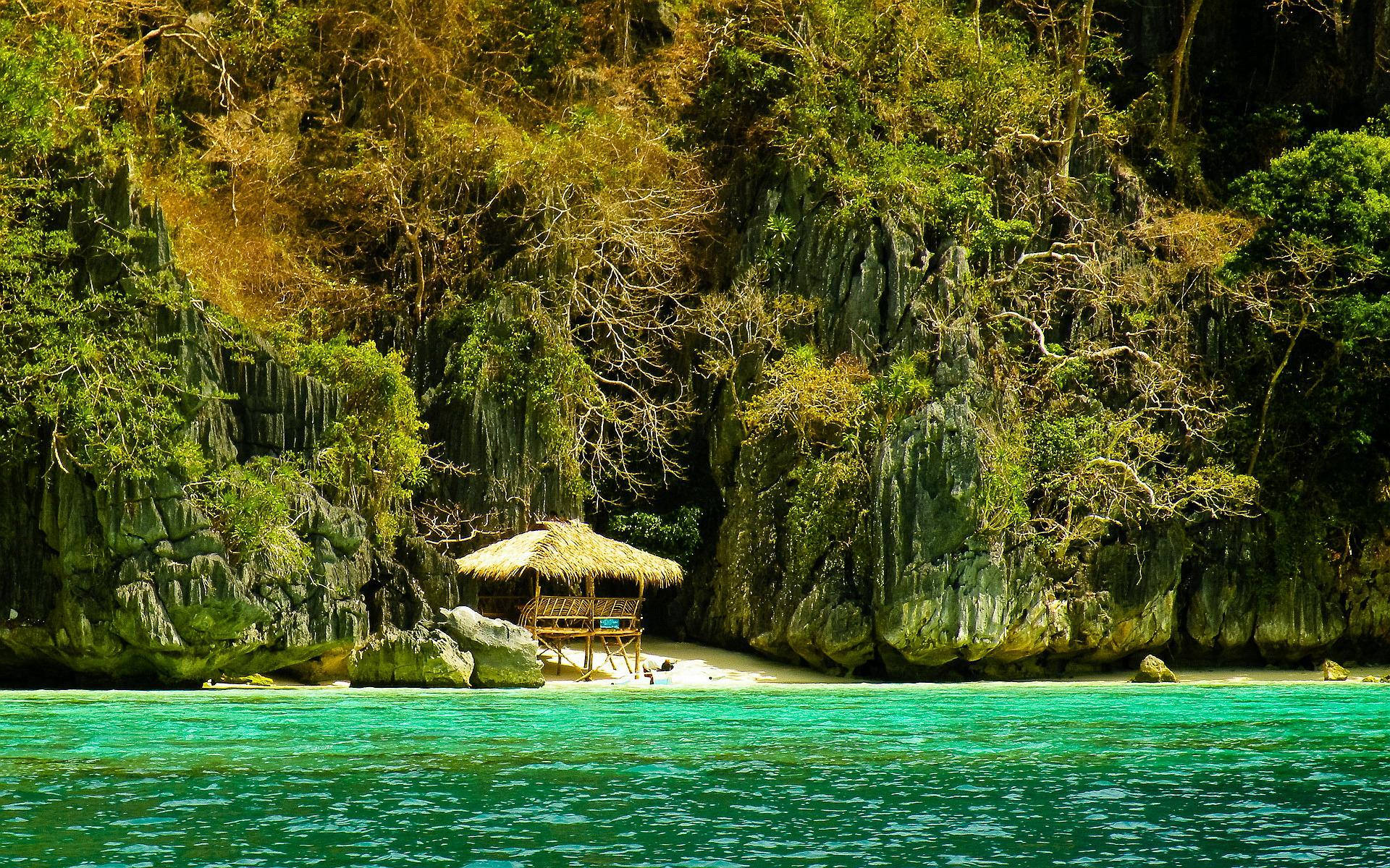 Philippines 桌布 壁紙 — 一間茅草屋頂的小屋坐落在白色沙灘上，旁邊是碧綠的海水，四周環繞著茂密的綠色懸崖和纏繞的樹木。