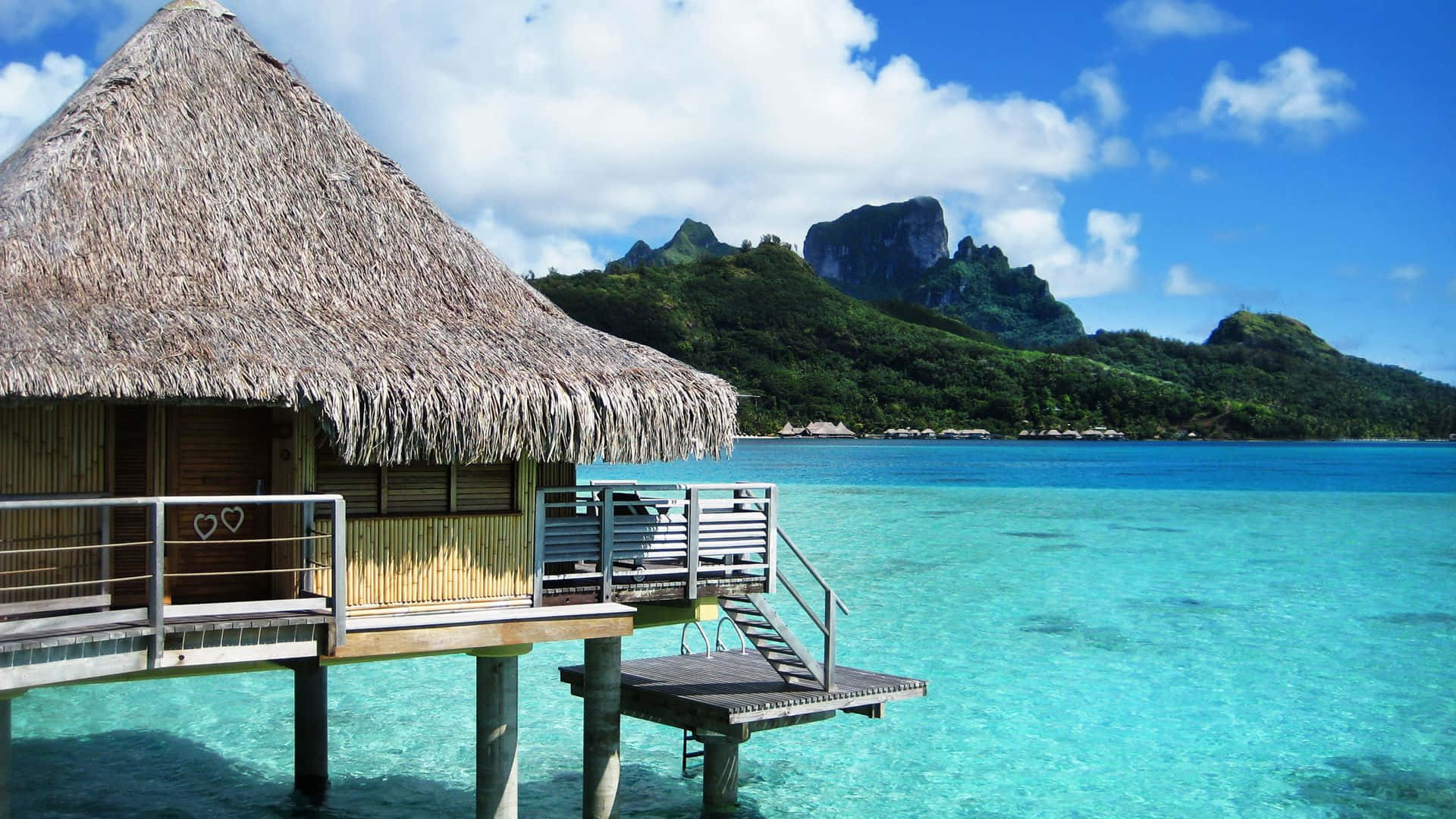 Bora Bora 桌布 壁紙 — 一間茅草屋頂的水上平房漂浮在清澈碧綠的海水上，鬱鬱蔥蔥的綠色山脈在明亮的藍天下聳立。