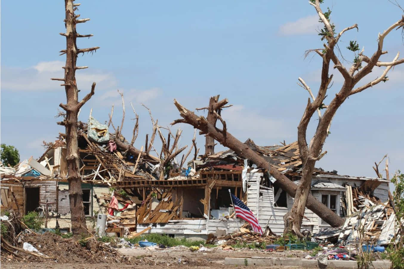 Natural Disaster 桌布 壁紙 — 一間被摧毀的房屋在晴朗的藍天下成為廢墟，樹木折斷，一面美國國旗仍在飄揚。