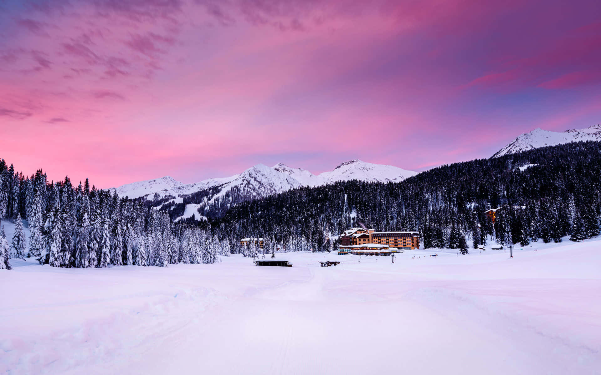 Ski Mountain 桌布 壁紙 — 一間被松樹環繞、覆蓋著白雪的山間小屋，在充滿活力的粉紅色和紫色日落天空下。