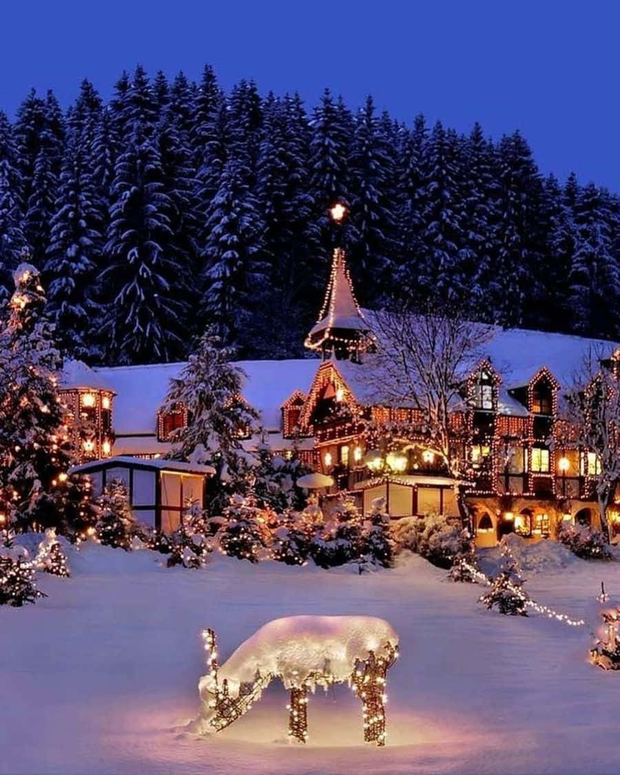 Christmas Winter Wonderland 桌布 壁紙 — 一間被雪覆蓋的小屋在暮色中溫暖地發光，前面有節日燈飾和發光的馴鹿雕塑。