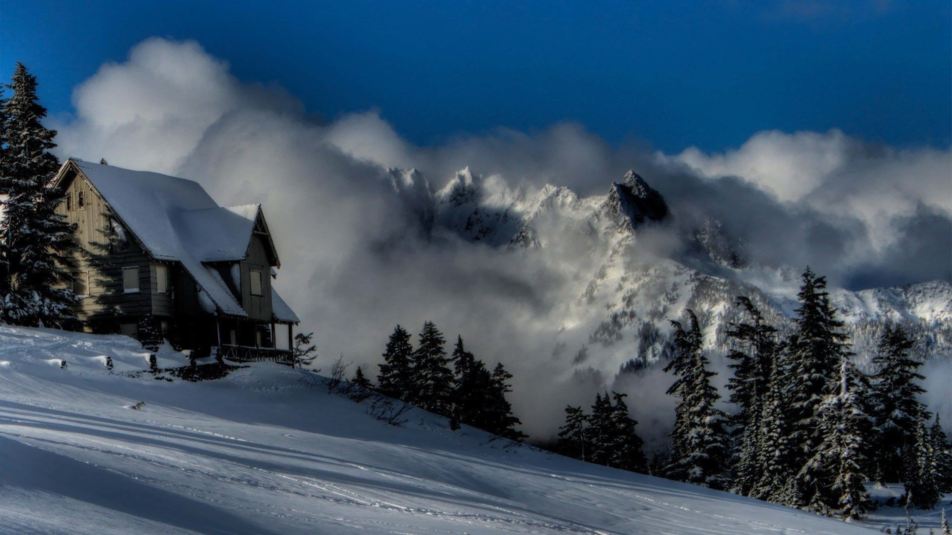 Snow 4K 桌布 壁紙 — 一間被雪覆蓋的小屋坐落在山坡上，雲朵在深藍色天空下圍繞著鋸齒狀的山峰旋轉。