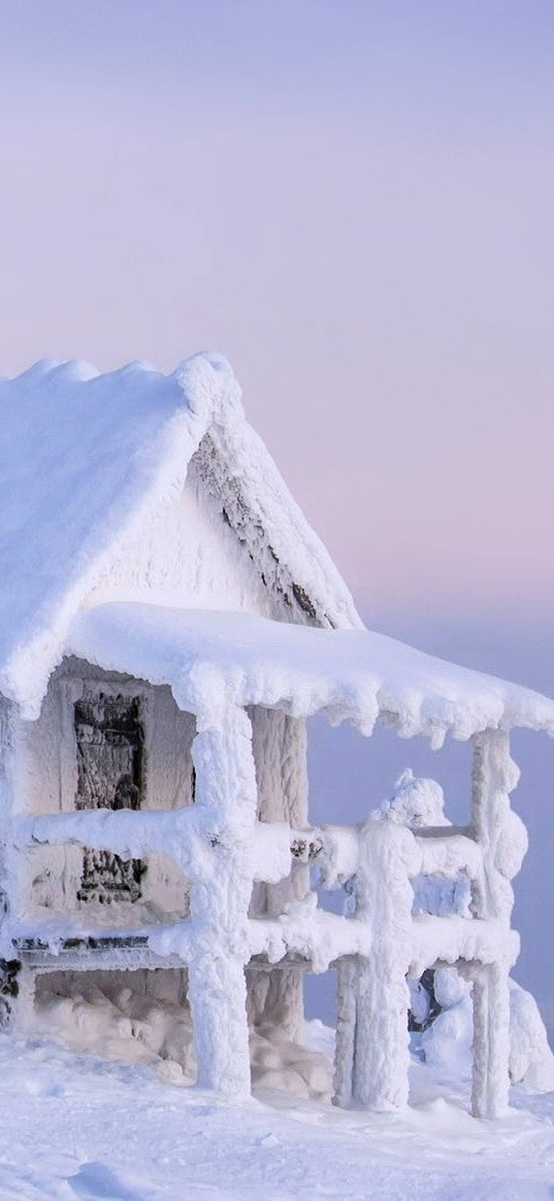 Cute Winter iPhone 桌布 壁紙 — 一間被雪覆蓋的木屋，屋頂掛著冰柱，背景是柔和的粉紅與藍色暮色天空。