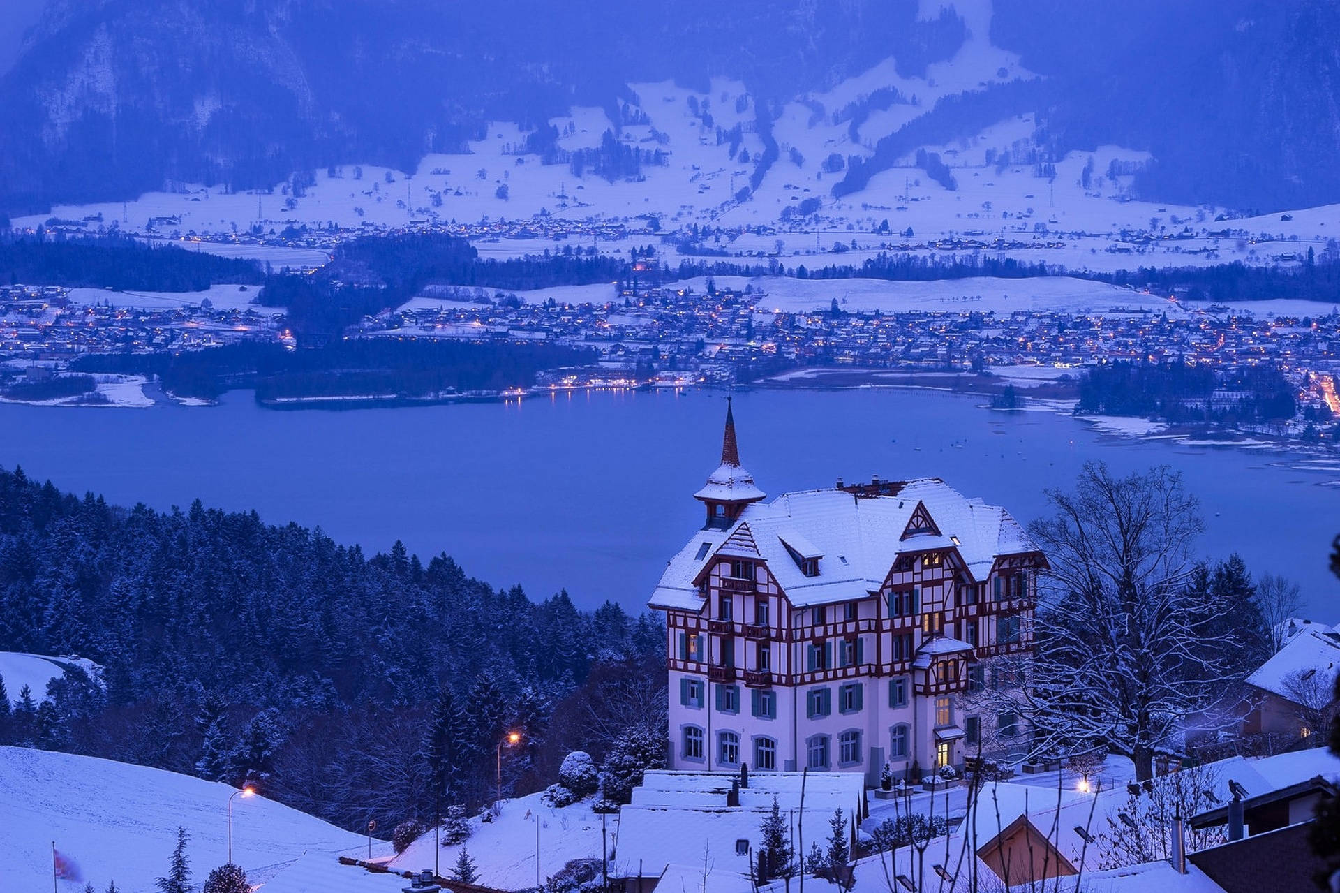 Alps 桌布 壁紙 — 一間被雪覆蓋的阿爾卑斯山小木屋在暮色中的湖泊和山脈背景下溫暖地發光。