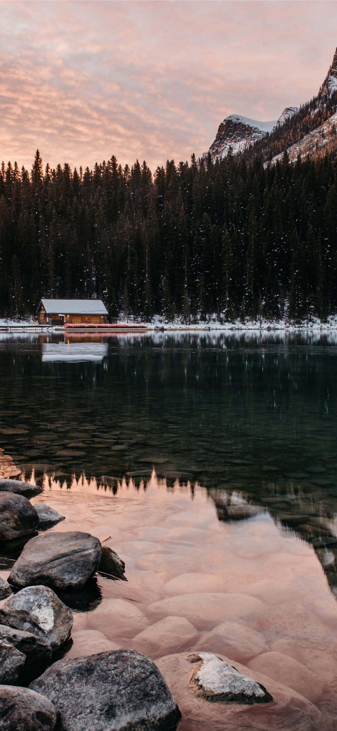 Cozy iPhone 桌布 壁紙 — 一間覆蓋著雪的小屋在黎明時分靜靜地坐落在結冰的湖面上，山脈和松樹倒映在平靜的水面上。