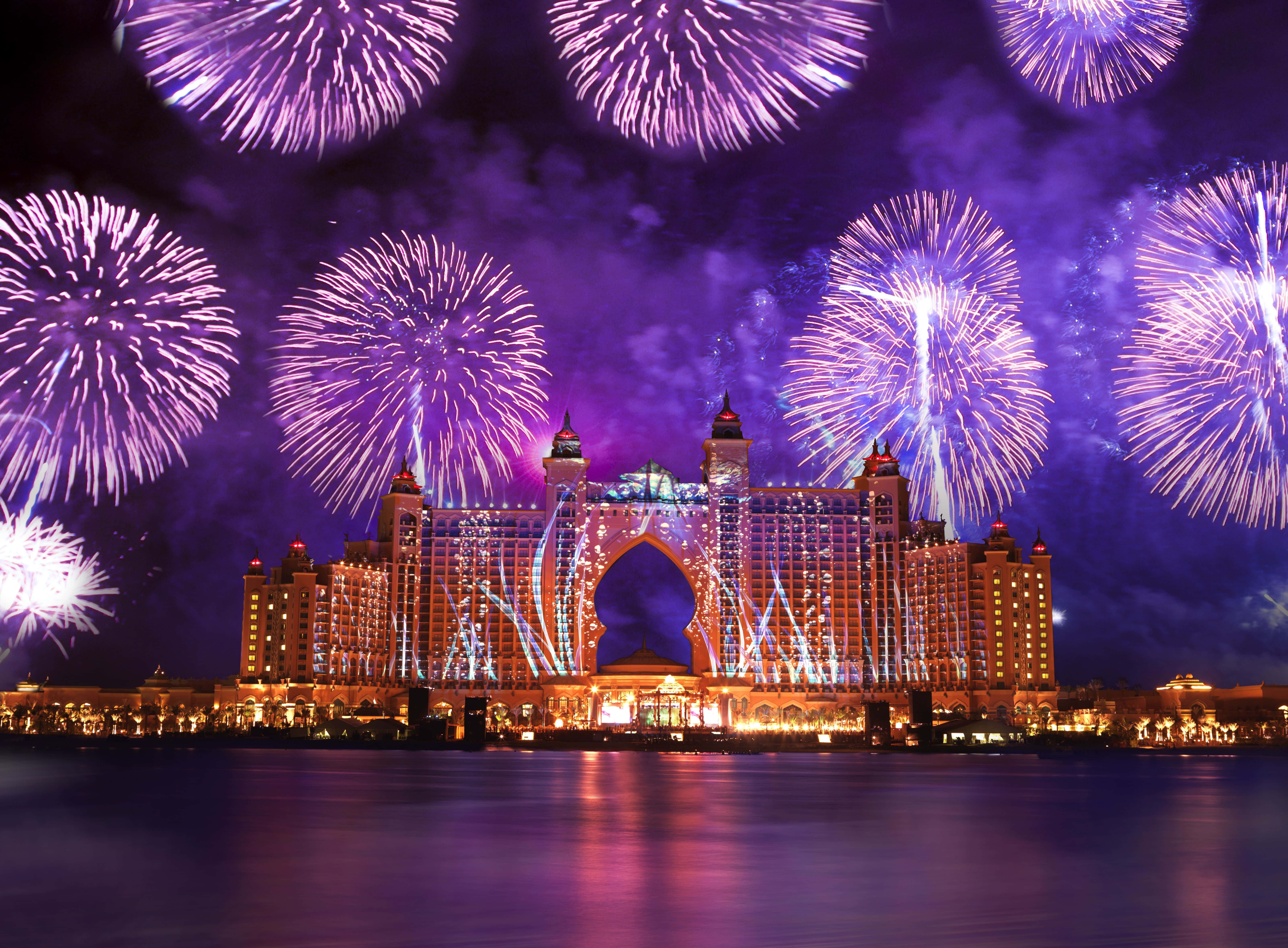 Fireworks 桌布 壁紙 — 一間豪華大酒店在夜晚亮起燈光，壯觀的紫色煙火在其標誌性拱門上方爆炸，並倒映在下方水面上。