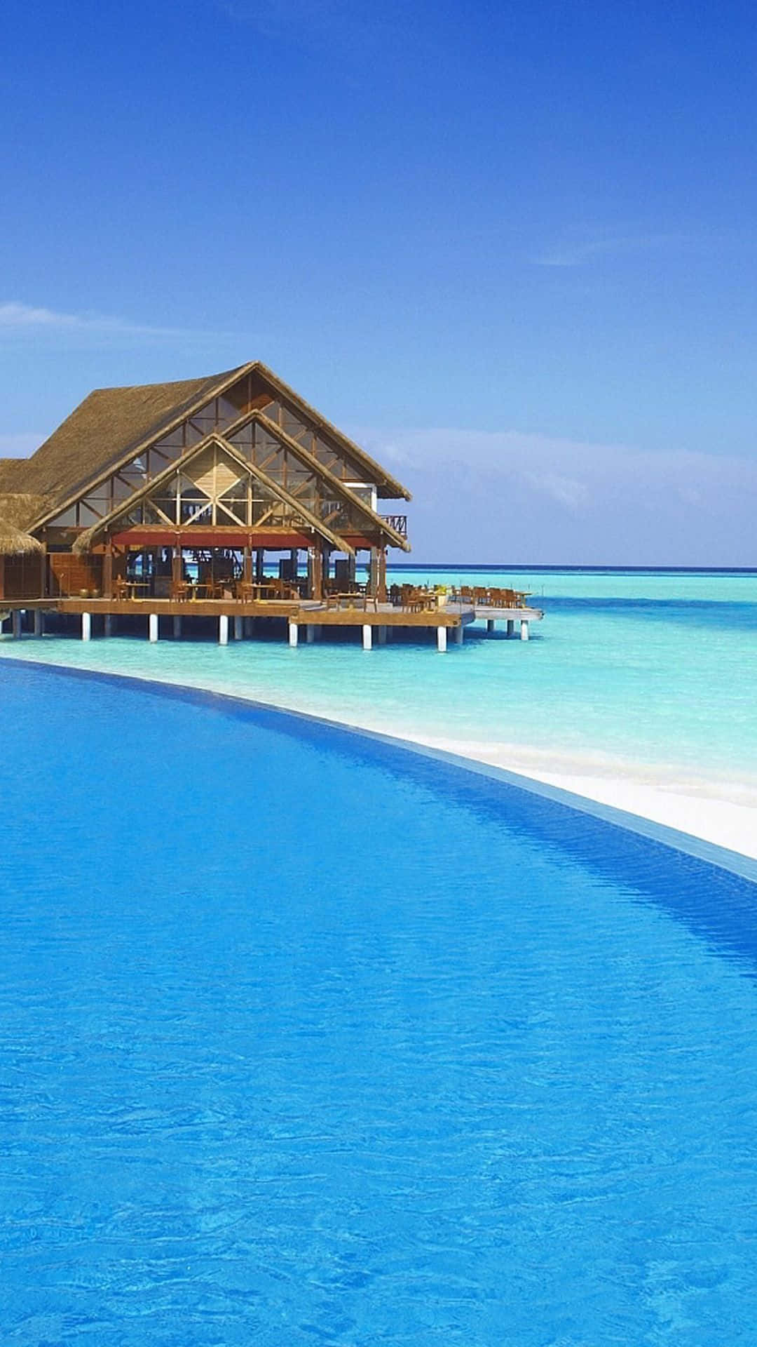 Beach Resort 桌布 壁紙 — 一間豪華的水上別墅，茅草屋頂，矗立在碧綠海水上的支柱上，旁邊是無邊際泳池，頭頂是清澈的藍天。