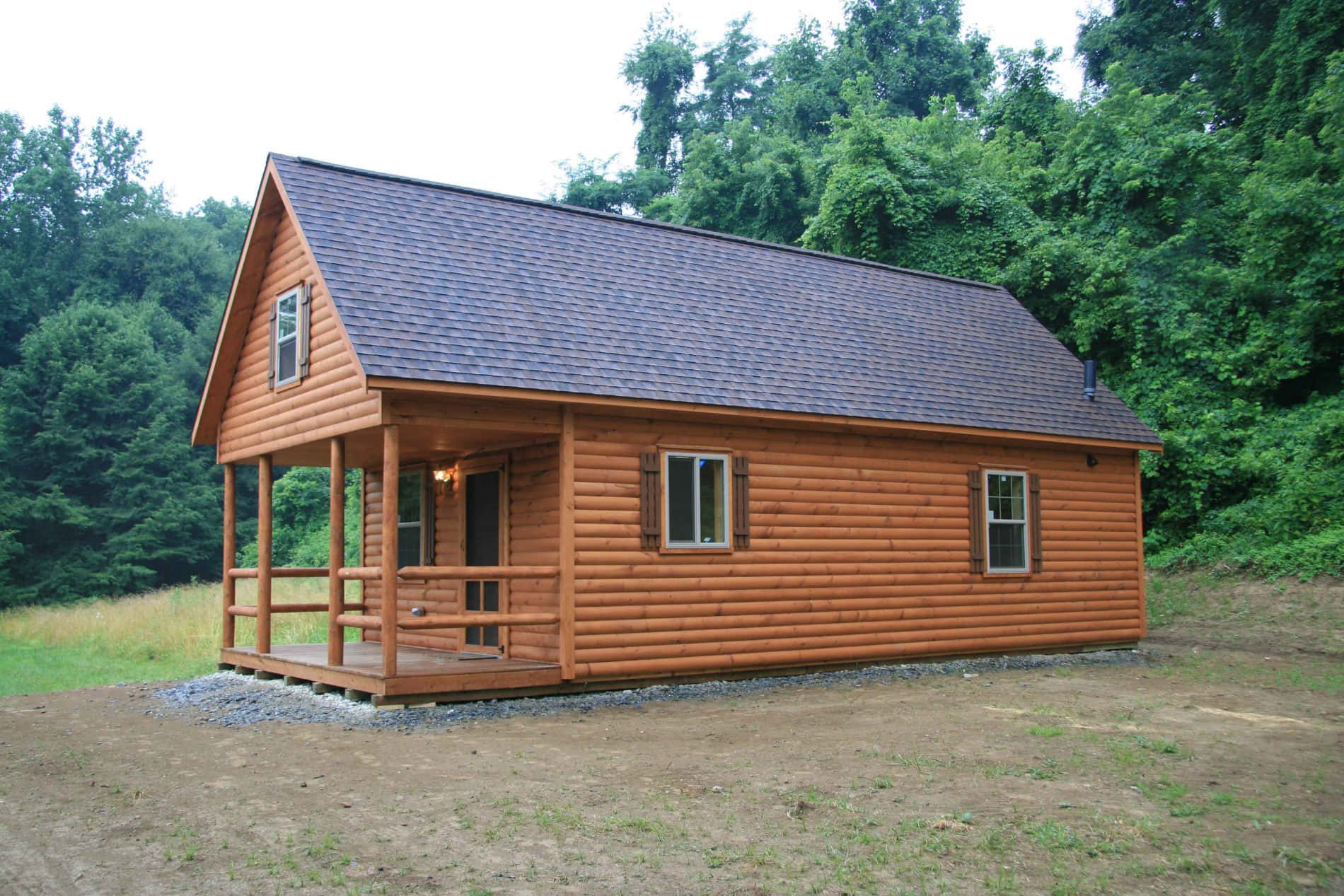 Log Cabin 桌布 壁紙 — 一間質樸的圓木小屋，深色屋頂和前門廊，坐落在碎石地上，周圍是茂密的綠色森林。
