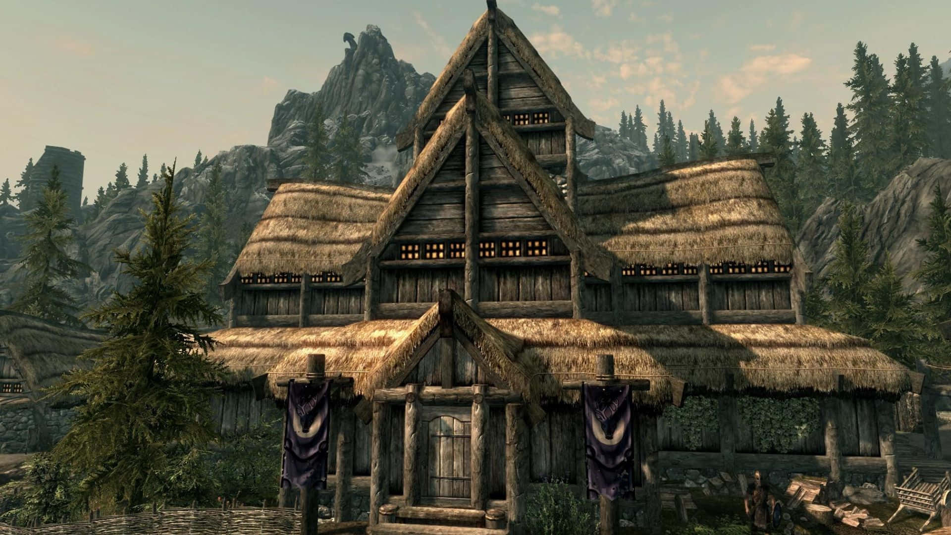 Skyrim Falkreath 桌布 壁紙 — 一間質樸的木屋，茅草屋頂和懸掛的橫幅，坐落在松樹和岩石山脈之間，天空柔和。