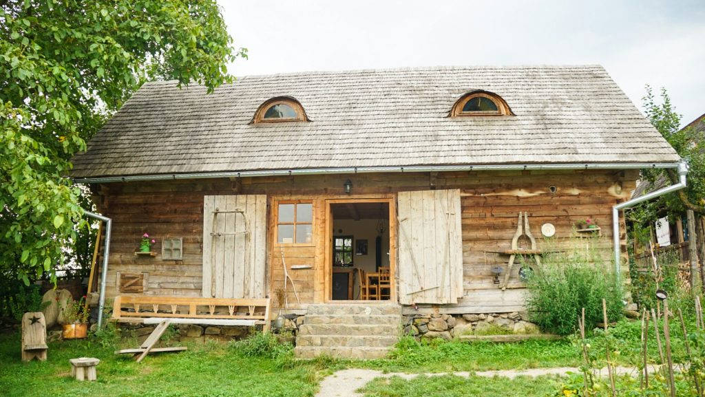 Romania 桌布 壁紙 — 一間質樸的木屋，配有風化的百葉窗和石頭地基，坐落在鬱鬱蔥蔥的花園中，天空多雲。