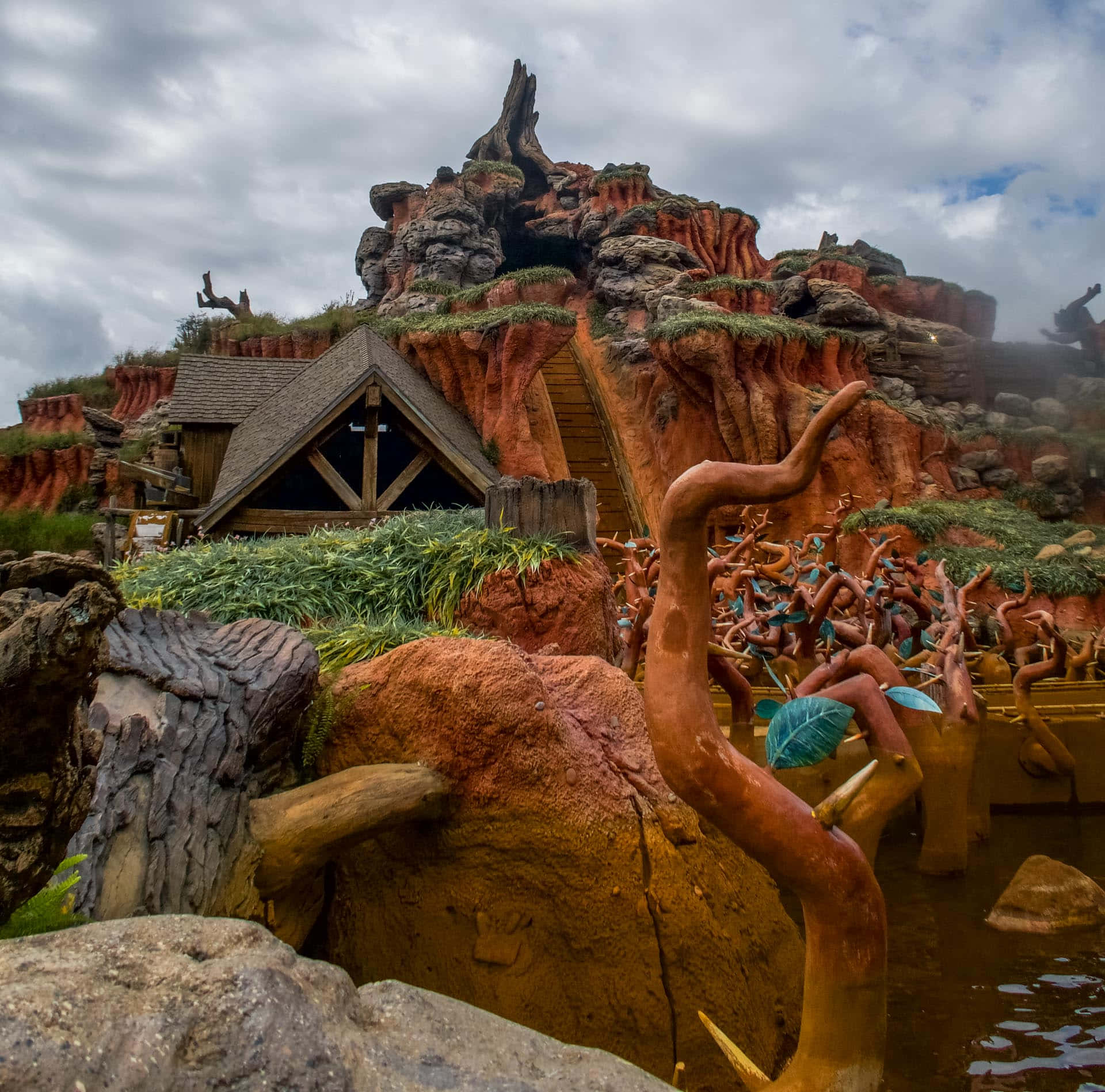 Splash Mountain 桌布 壁紙 — 一間質樸的木屋坐落在一個夢幻般的紅岩峽谷中，有扭曲的樹根和長滿苔蘚的懸崖，天空多雲。