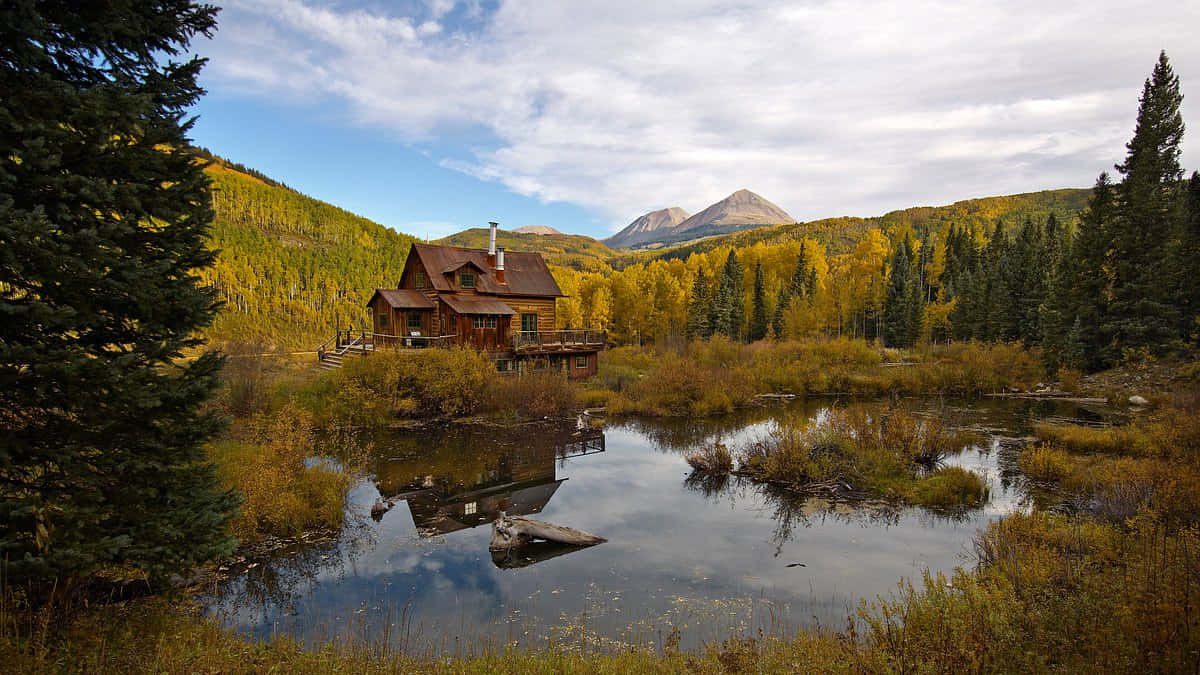 Fall Cabin 桌布 壁紙 — 一間質樸的木屋坐落在平靜的池塘邊，池塘倒映著山脈，天空是部分多雲的秋日天空。