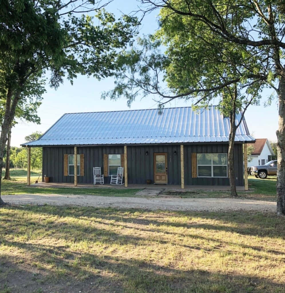 房屋 桌布 壁紙 — 一間質樸的金屬屋頂小屋，配有木柱和搖椅，在陽光明媚的日子裡坐落在枝葉繁茂的樹下。