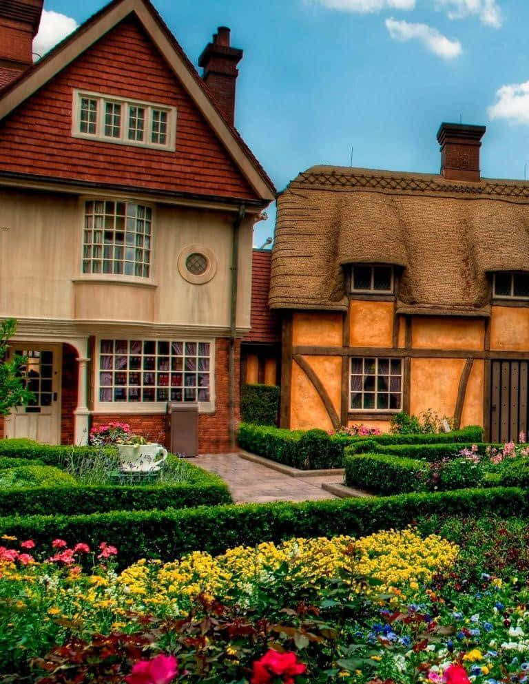 Beautiful House 桌布 壁紙 — 一間迷人的小屋，擁有紅色木瓦和茅草屋頂，坐落在充滿活力的花園中，花園裡開滿了色彩繽紛的花朵，上方是清澈的藍天。
