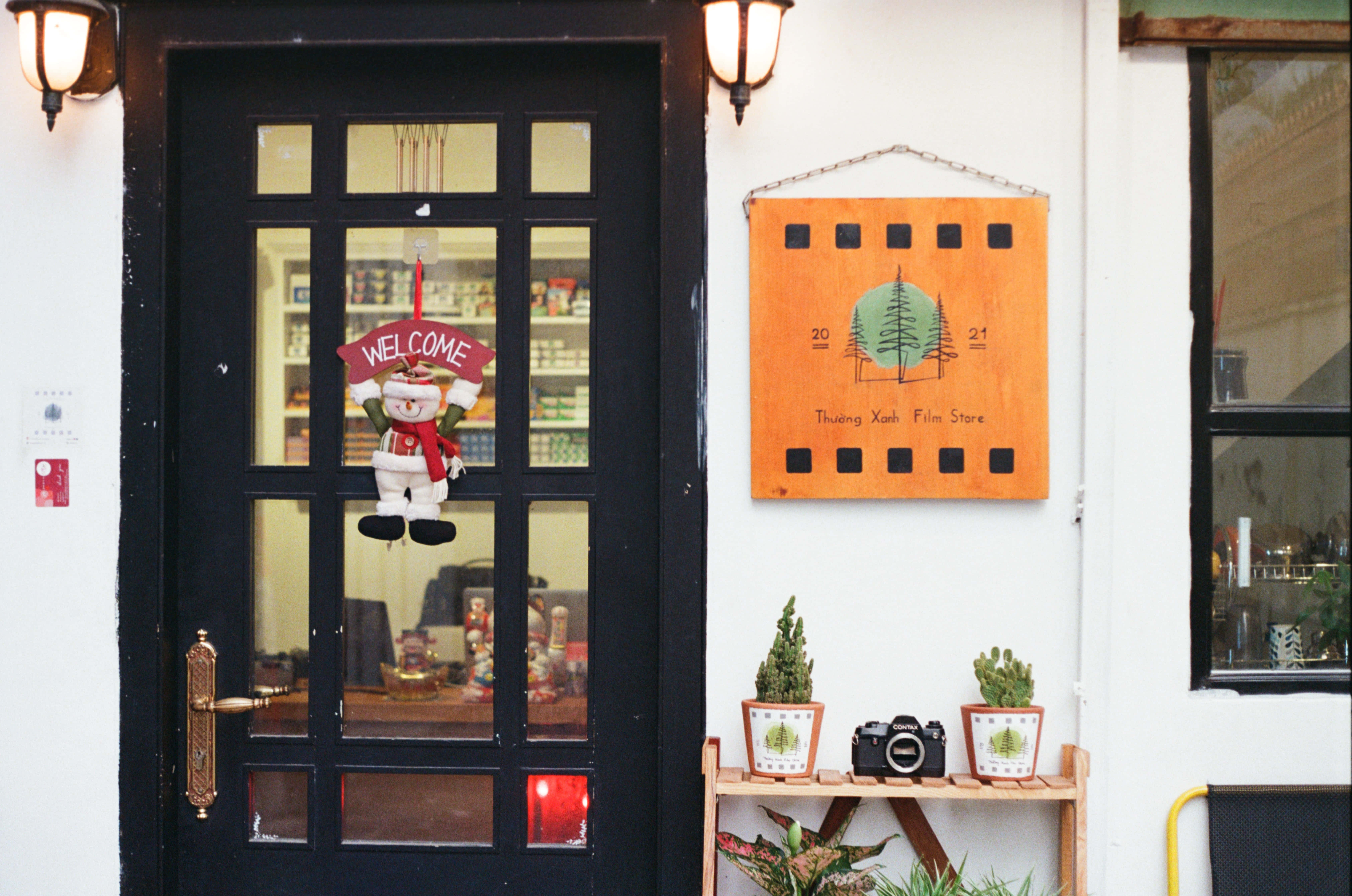 Welcome 桌布 壁紙 — 一間迷人的底片店入口，黑色門上掛著雪人歡迎牌，植物和相機的架子上方有一個木製底片捲牌。