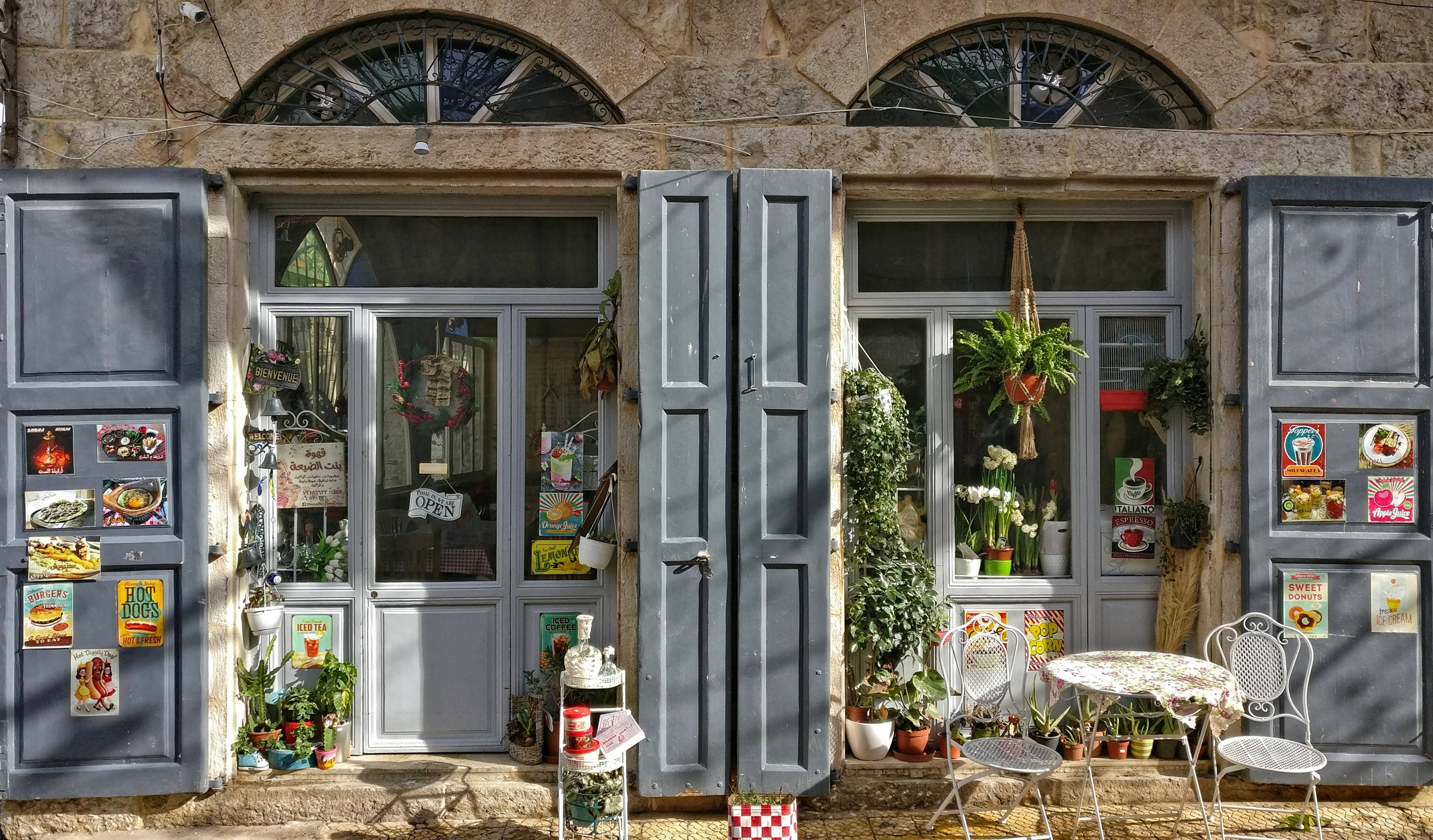 Lebanon 桌布 壁紙 — 一間迷人的石牆咖啡館，配有藍色百葉窗和懸掛植物，沐浴在溫暖的陽光中。