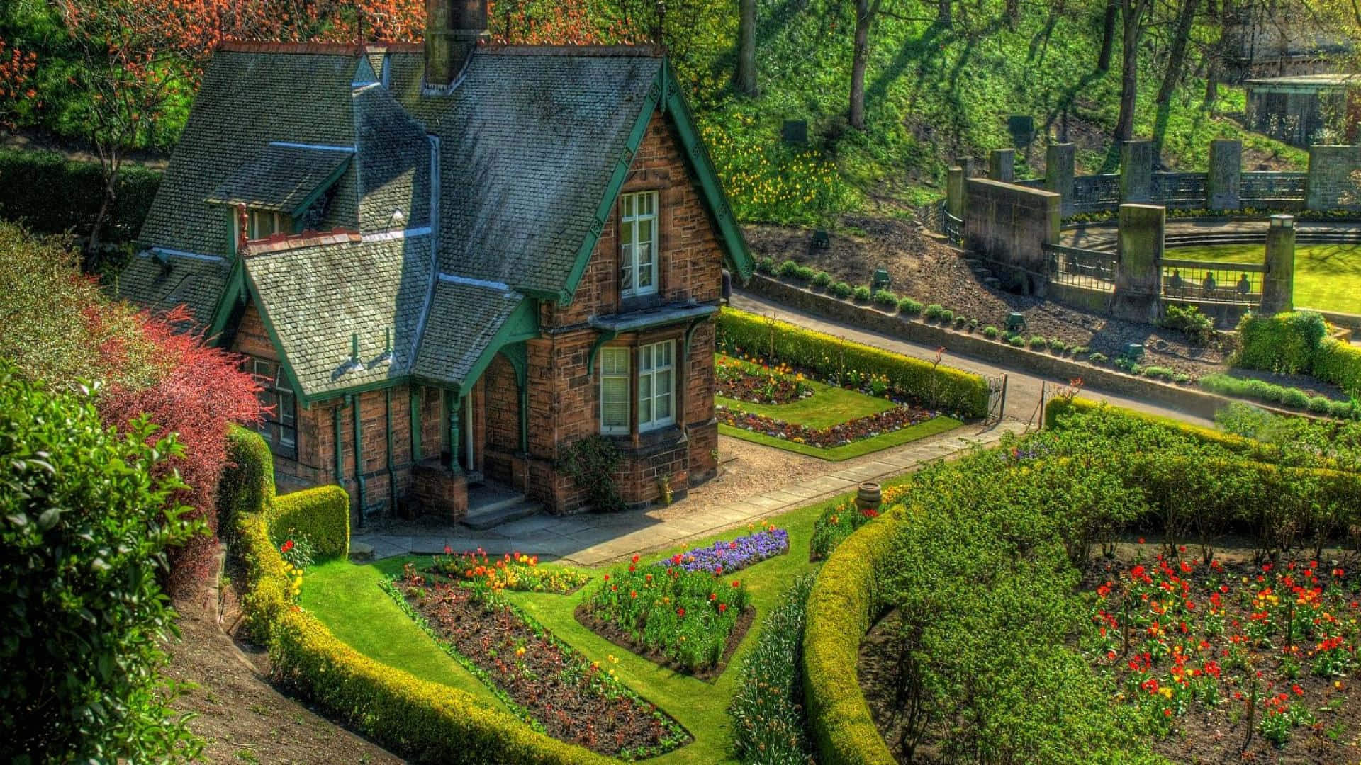 Cottage Garden 桌布 壁紙 — 一間迷人的石頭小屋，帶有綠色飾邊，坐落在充滿活力的花園中，花園裡有色彩繽紛的花朵和修剪整齊的樹籬，沐浴在明亮的陽光下。