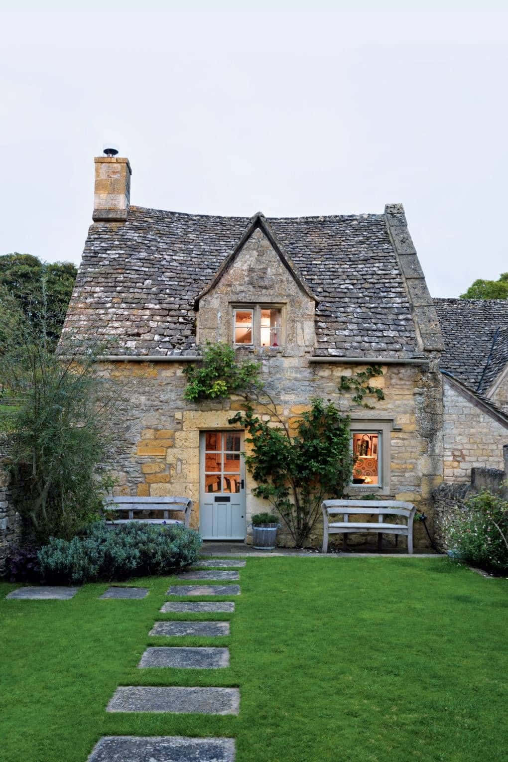 Cottage 桌布 壁紙 — 一間迷人的石頭小屋，配備石板屋頂和發光的窗戶，坐落在鬱鬱蔥蔥的綠色花園中，有石徑和木製長椅。