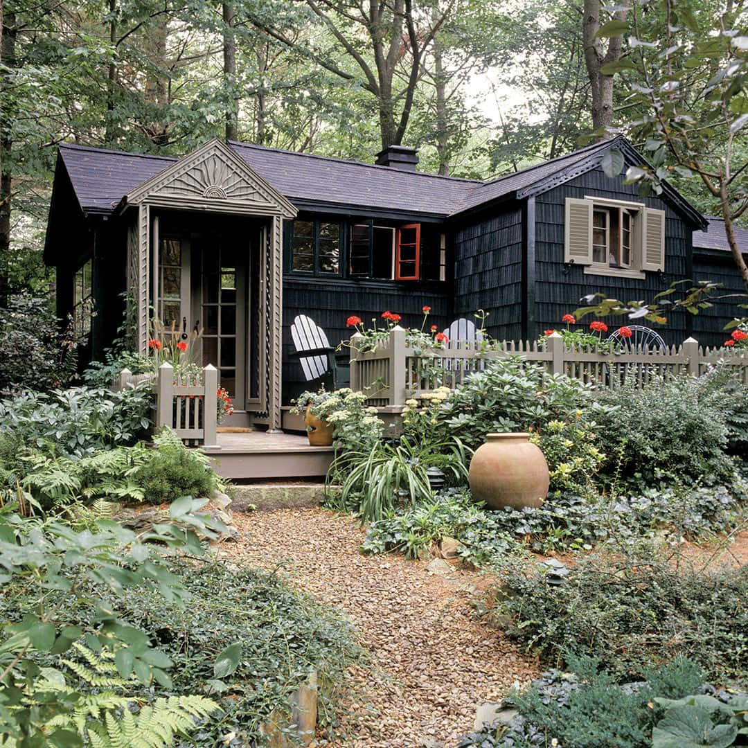 Cottage 桌布 壁紙 — 一間迷人的黑色木瓦小屋，坐落在鬱鬱蔥蔥的綠色森林中，一條碎石路通向點綴著紅花和白椅的門廊。
