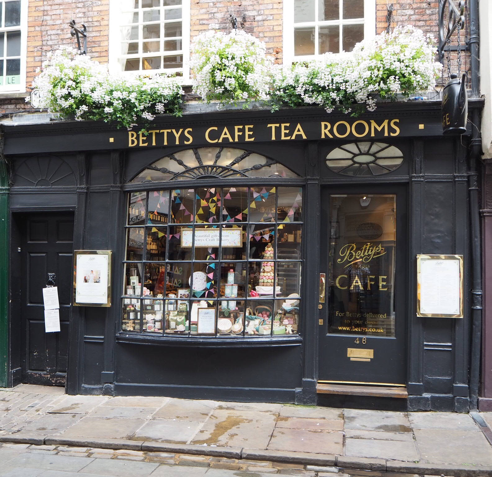 England 桌布 壁紙 — 一間迷人的黑色茶室，窗台上的白色花朵盛開，櫥窗內掛滿色彩繽紛的彩旗。