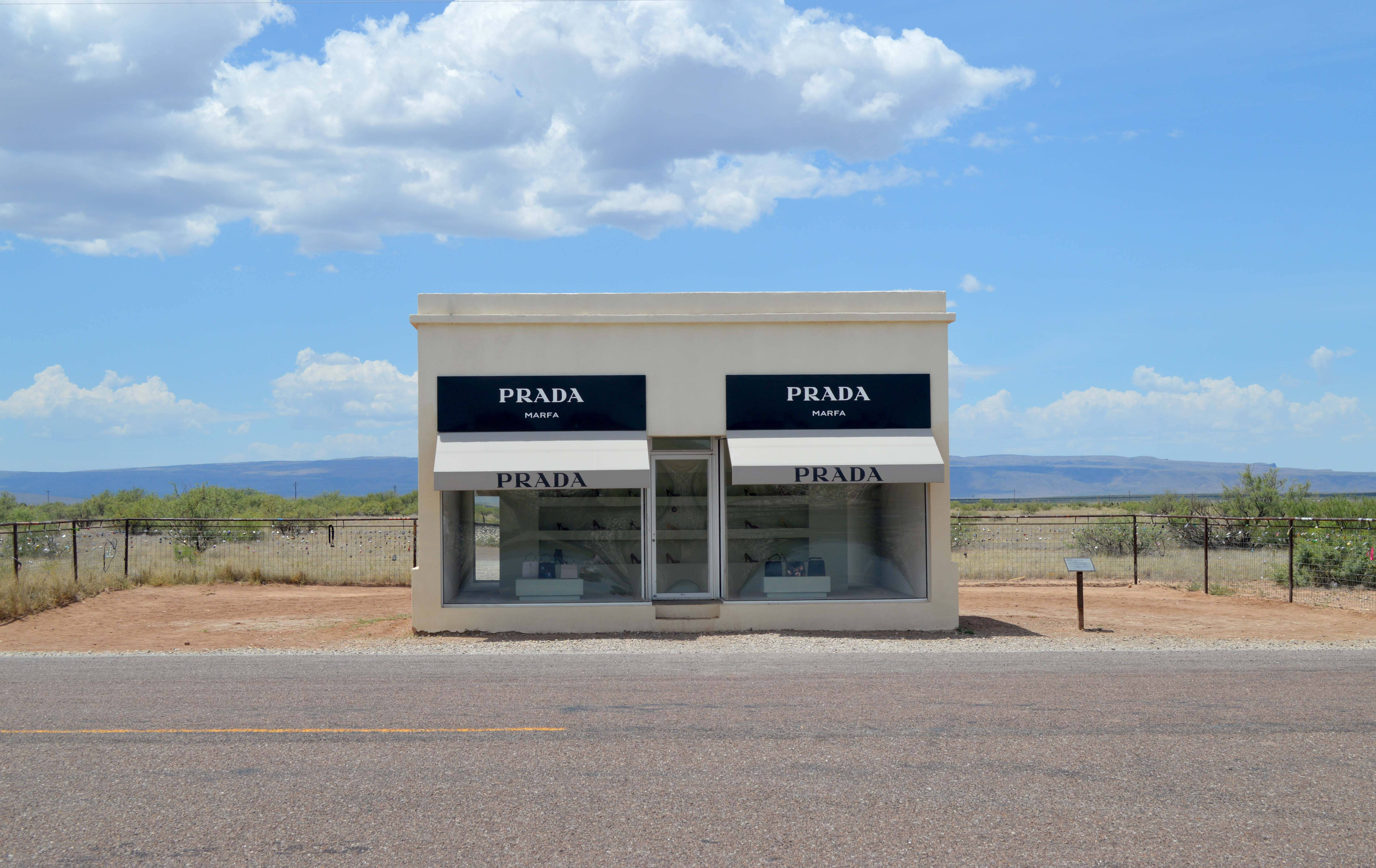 Prada 桌布 壁紙 — 一間醒目的Prada商店獨自矗立在廣闊的沙漠景觀中，上方是明亮的藍天和蓬鬆的雲朵。