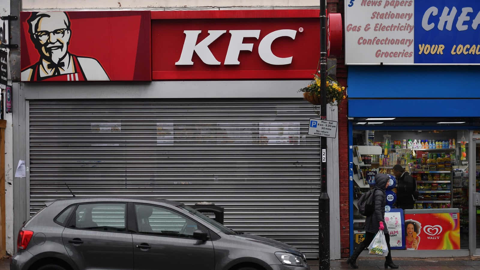 肯德基 桌布 壁紙 — 一間關閉的KFC店面，裝有金屬捲簾，旁邊有一間便利商店和一輛經過的汽車，位於城市街道上。