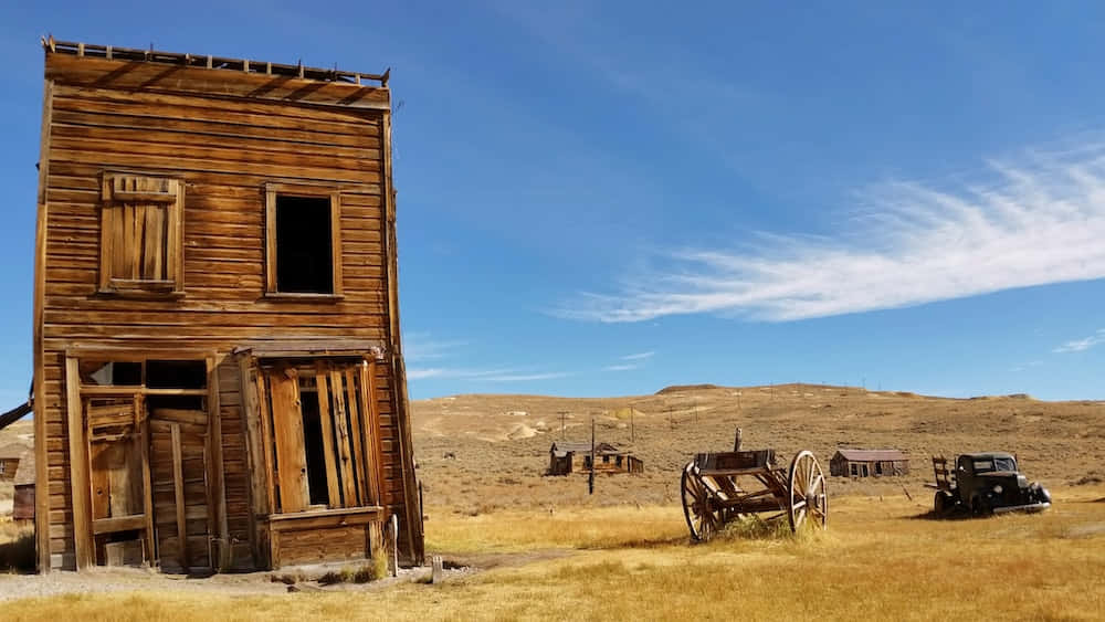 Old Western 桌布 壁紙 — 一間風化的木製沙龍被遺棄在烈日曝曬的沙漠中，附近有一輛破舊的馬車和一輛舊車，在清澈的藍天下。